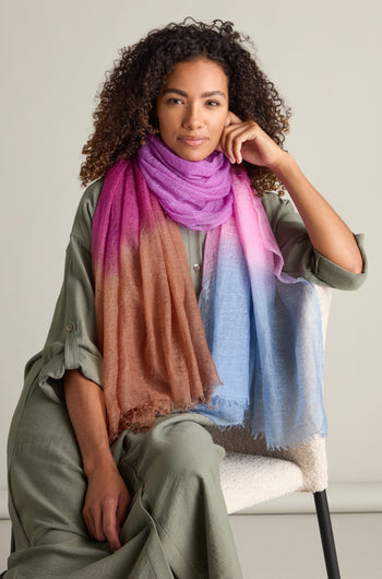 A person with curly hair wearing a green outfit and a St Tropez Cashmere Shawl, featuring a gradient watercolour design, is sitting on a white chair against a plain background.