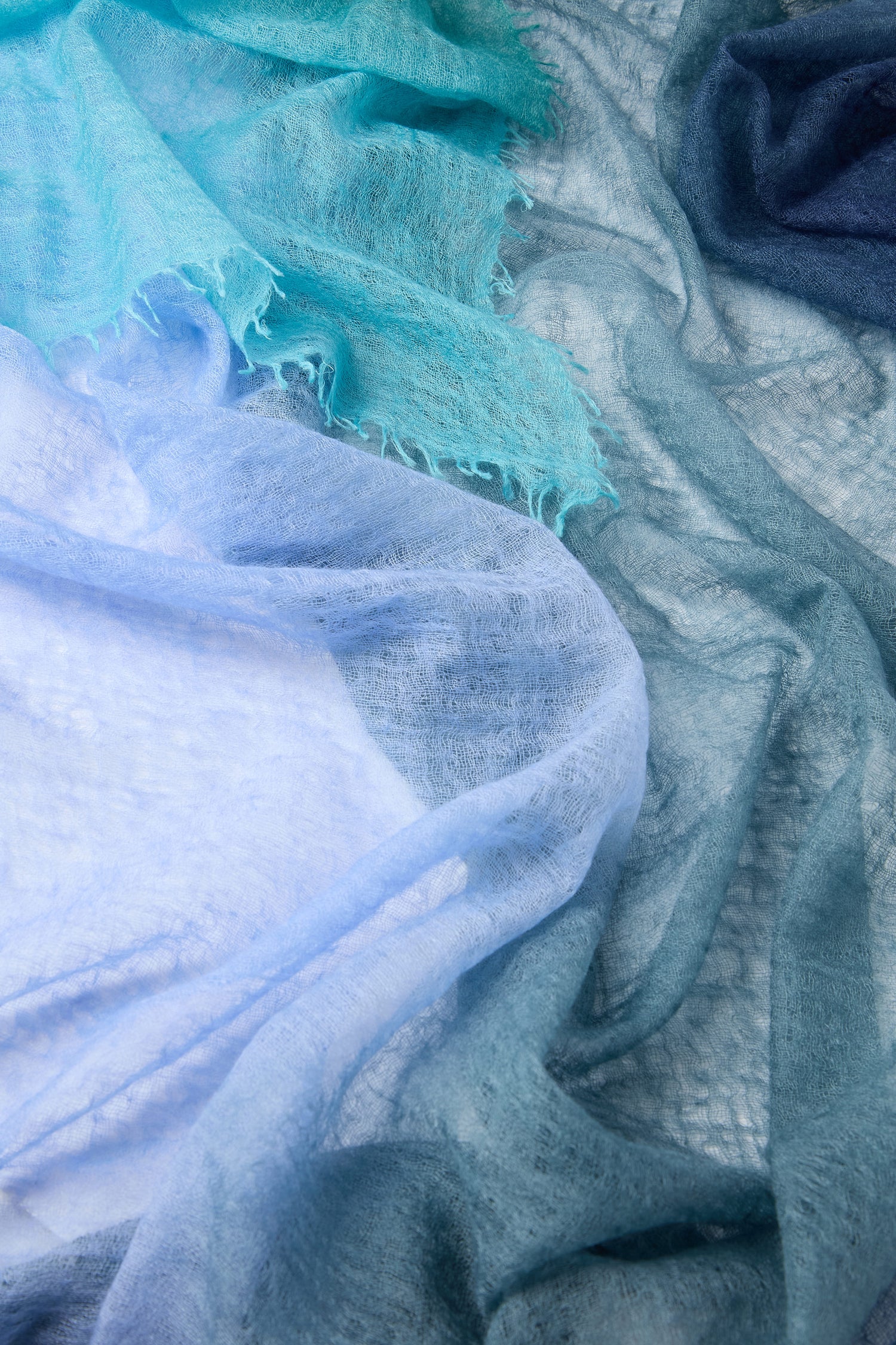 Close-up image of the textured, lightweight St Tropez Cashmere Shawl in shades of blue and teal layered on a flat surface, resembling a gradient watercolor design.