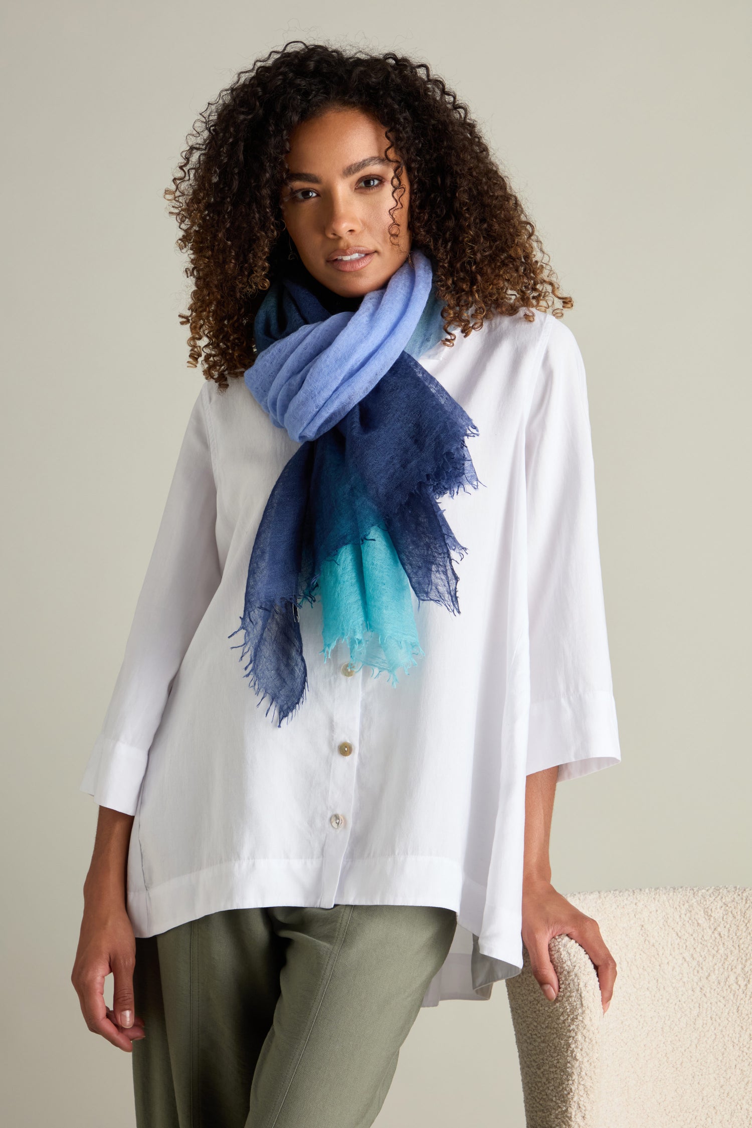 A person stands wearing a white blouse, green pants, and a luxurious accessory—a blue gradient watercolour design St Tropez Cashmere Shawl.