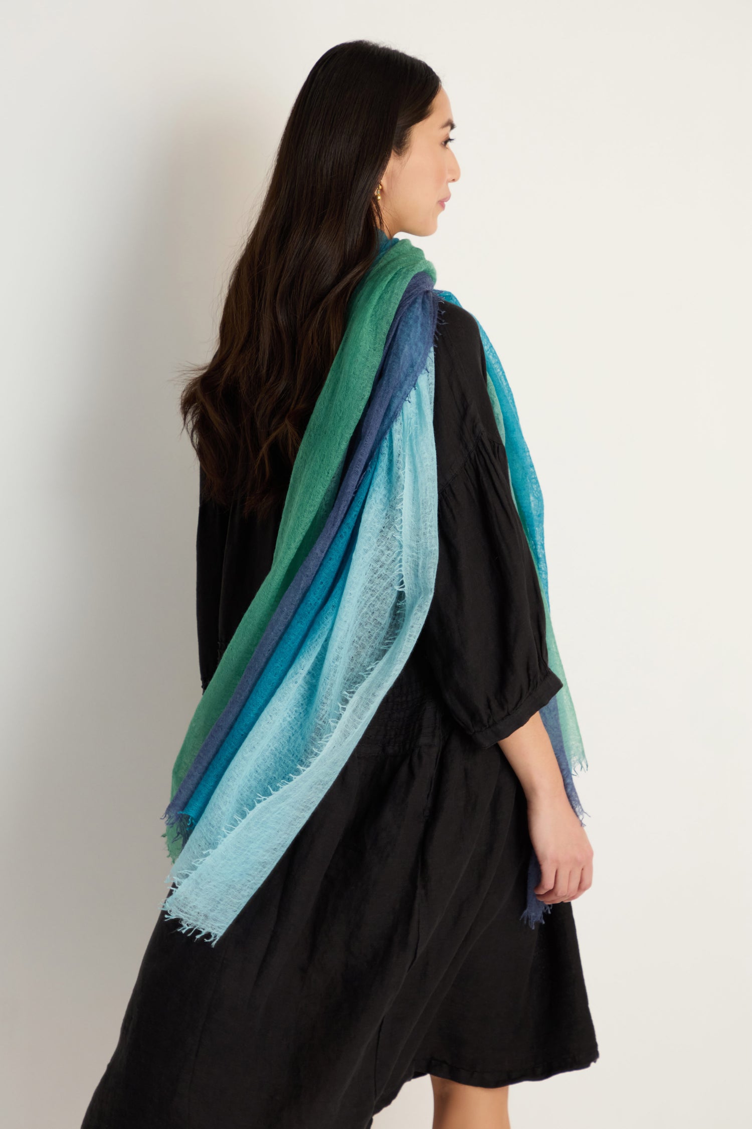 A woman in a black dress stands sideways, elegantly draping the St. Tropez Cashmere Shawl in green and blue tones—ideal for lightweight layering—against a plain white background, capturing effortless style.