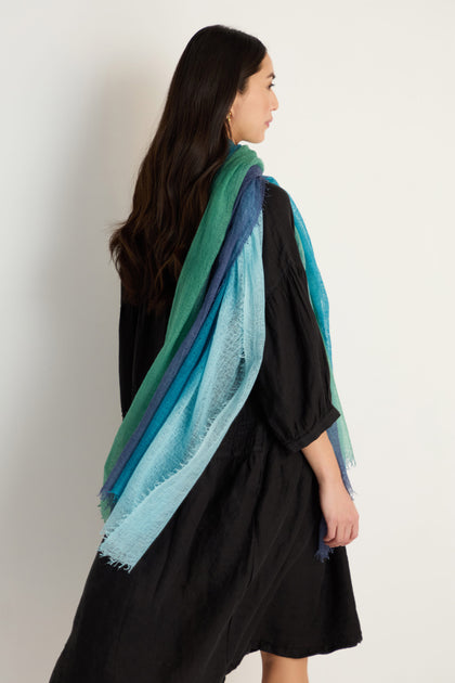 A woman in a black dress stands sideways, elegantly draping the St. Tropez Cashmere Shawl in green and blue tones—ideal for lightweight layering—against a plain white background, capturing effortless style.
