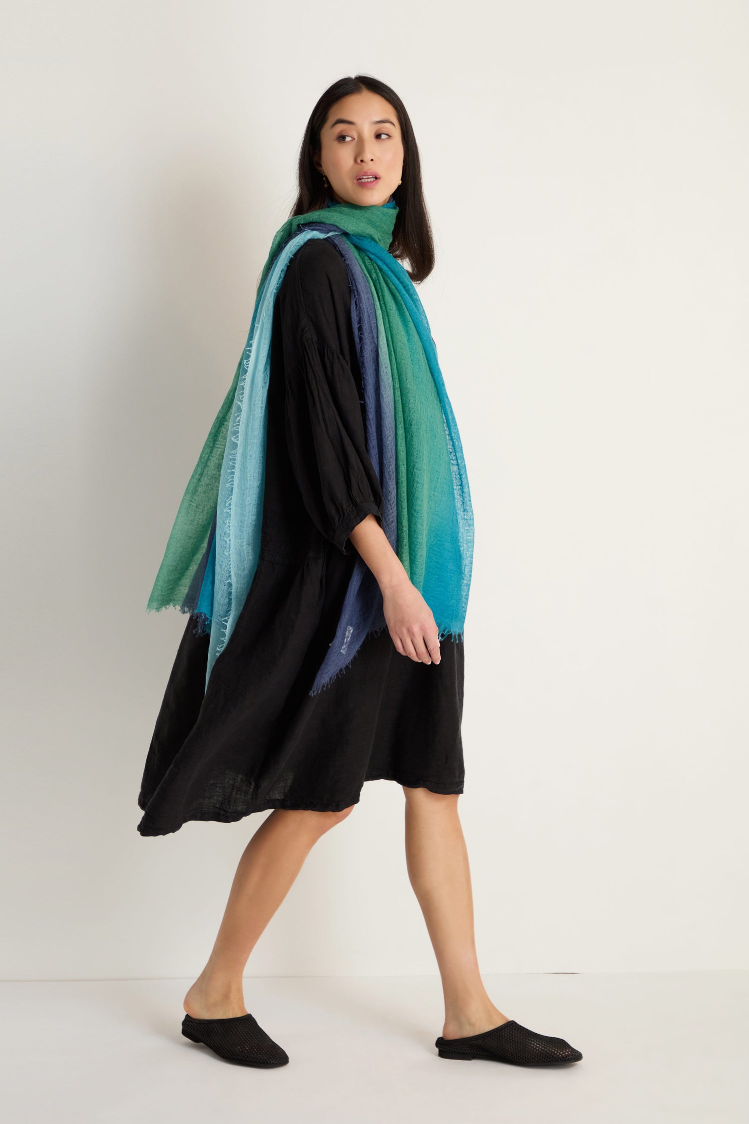 A woman in a black dress and shoes walks with the St. Tropez Cashmere Shawl, its lightweight gradient blue and green hues evoking elegance, draped over her shoulders against a plain background.