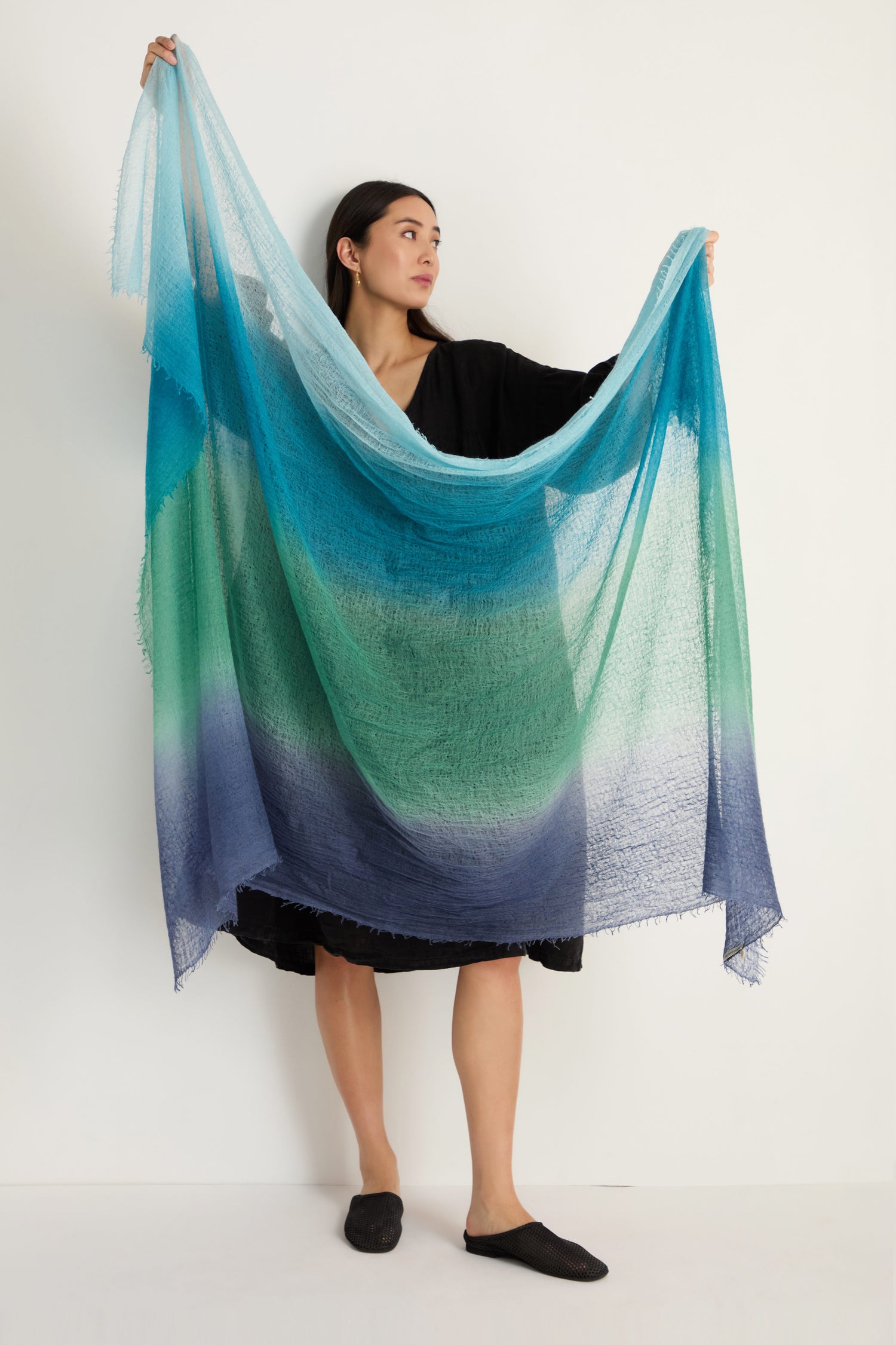 A woman in a black dress displays the St. Tropez Cashmere Shawl, featuring a large, semi-transparent design with gradient blue and green hues—ideal for lightweight layering against a plain backdrop.