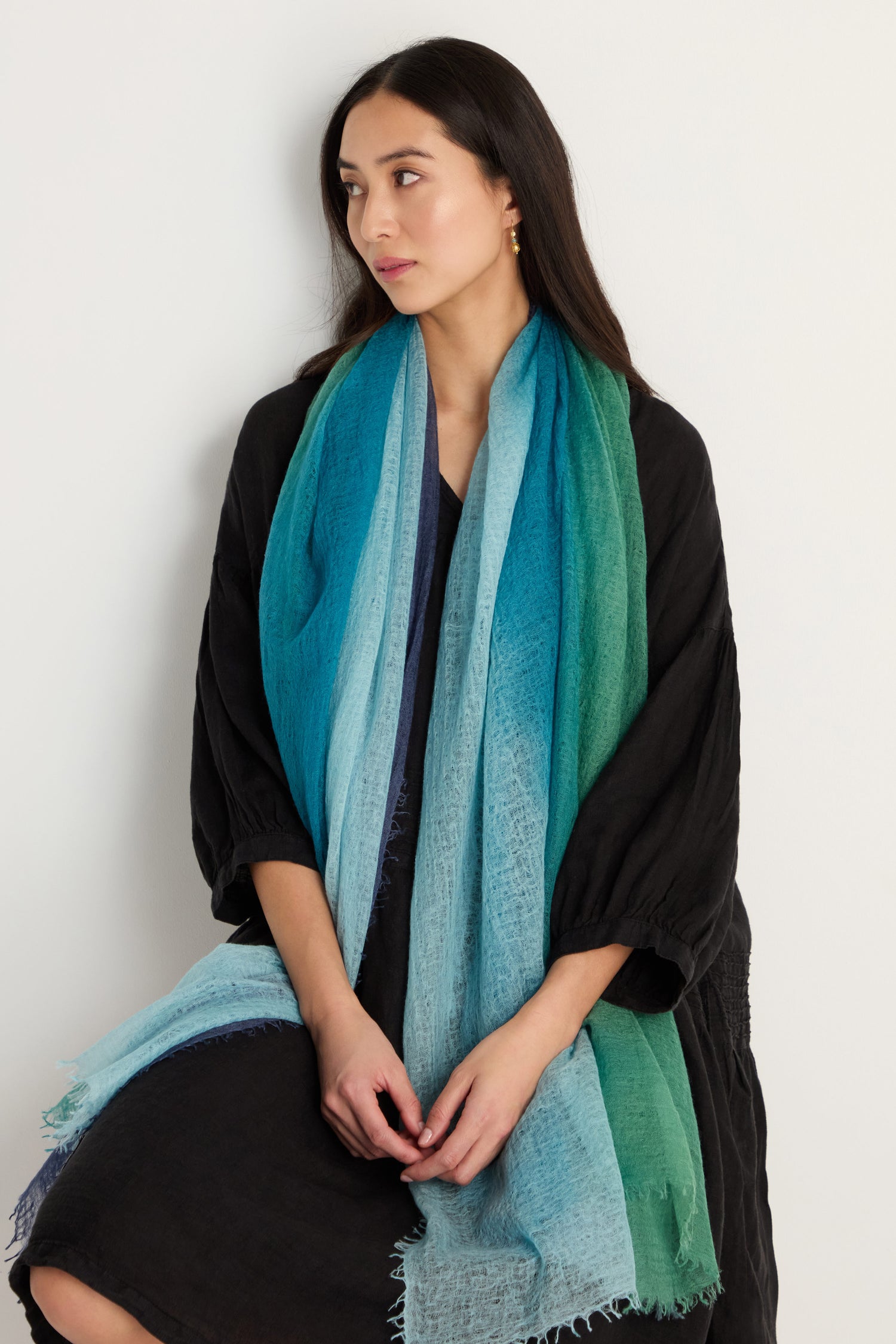 A woman with long dark hair, in a dark dress, wears the St. Tropez Cashmere Shawl—a large blue and green gradient wrap—while seated against a plain light-colored background.