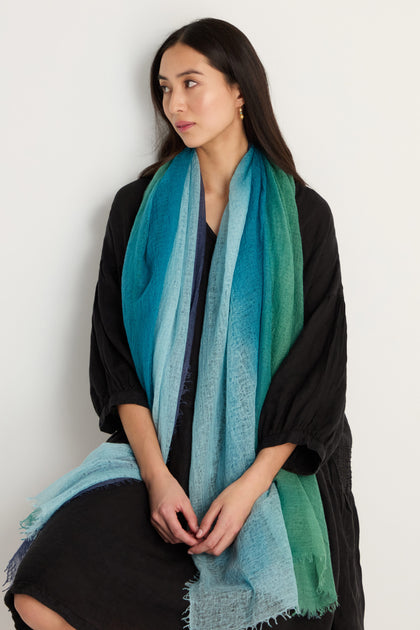 A woman with long dark hair, in a dark dress, wears the St. Tropez Cashmere Shawl—a large blue and green gradient wrap—while seated against a plain light-colored background.