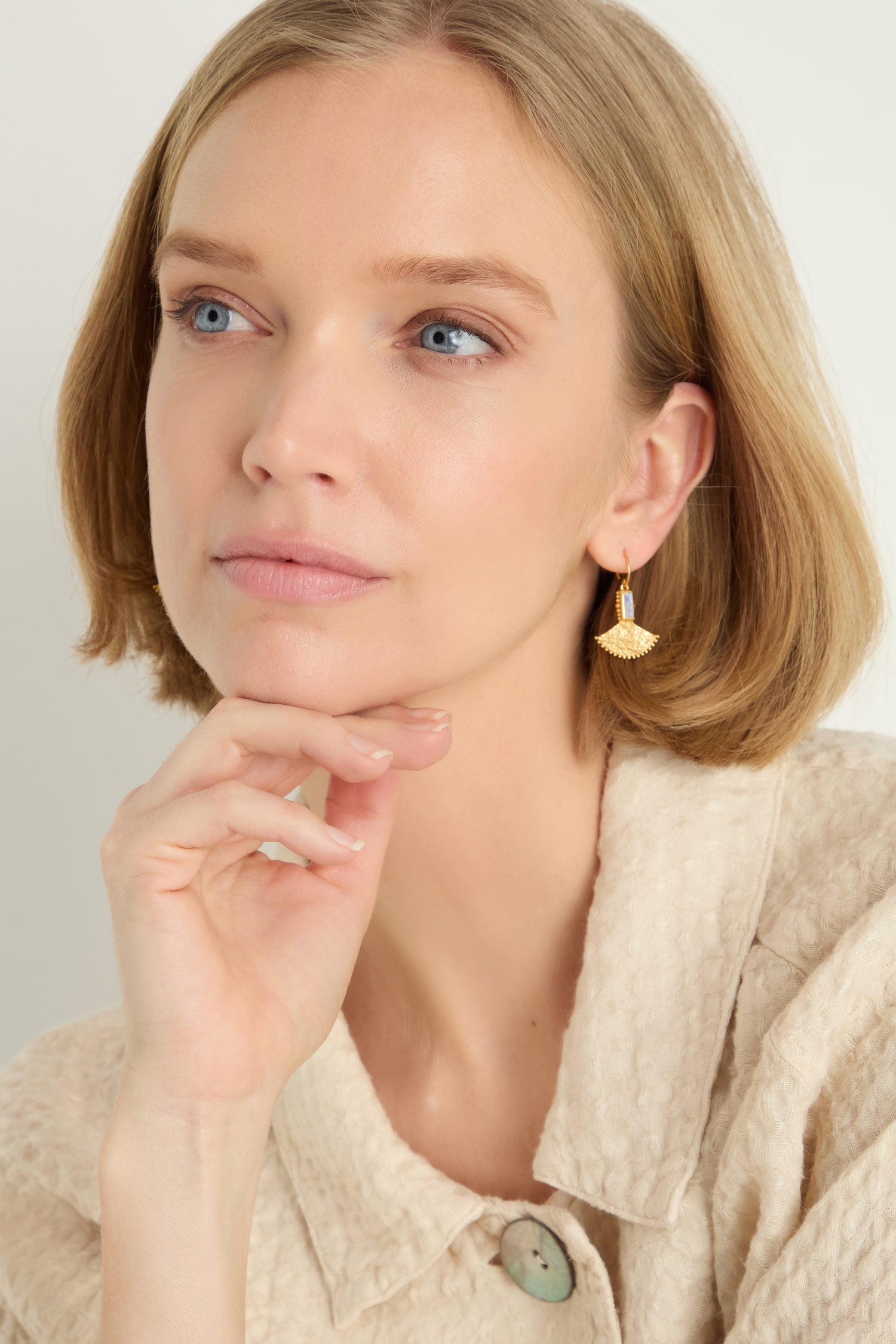 A woman with short blonde hair wears a textured beige top and the Gold Plated White Moonstone Mini Fan Earrings, resting her chin on her hand and gazing slightly upward.