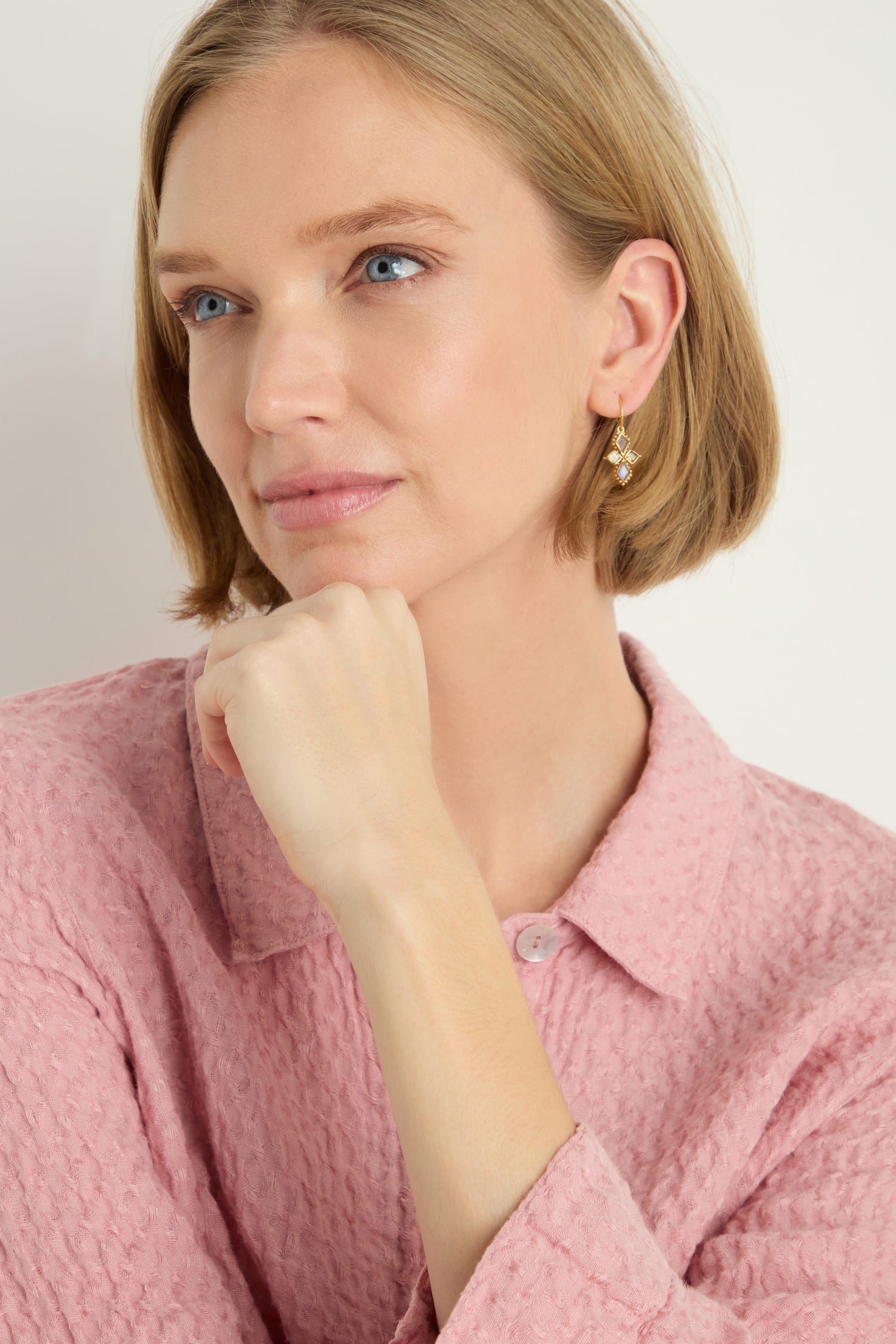 A woman with short blonde hair in a pink textured shirt rests her chin on her hand, looking thoughtfully to the side. She is wearing Gold Plated White Moonstone Kite Earrings.