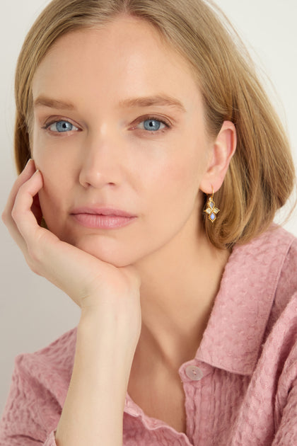 A woman with short blonde hair wears a textured pink shirt and Gold Plated White Moonstone Kite Earrings, resting her chin on her hand and looking at the camera.