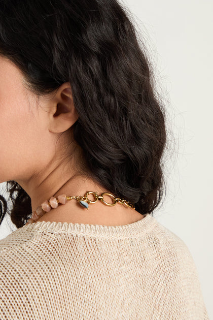 A woman with dark wavy hair, seen from the back and side, wears a beige knit top and the Peach Moonstone Bead Necklace—featuring gold chain, faceted beads, pink stones, and a blue accent.
