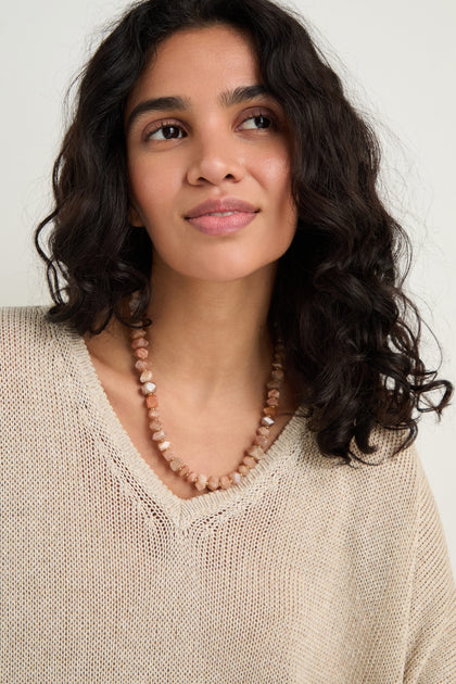Woman with wavy dark hair wears a beige knit sweater and the Peach Moonstone Bead Necklace, featuring faceted beads and a gold toggle clasp, as she gazes slightly to the side with a neutral expression.