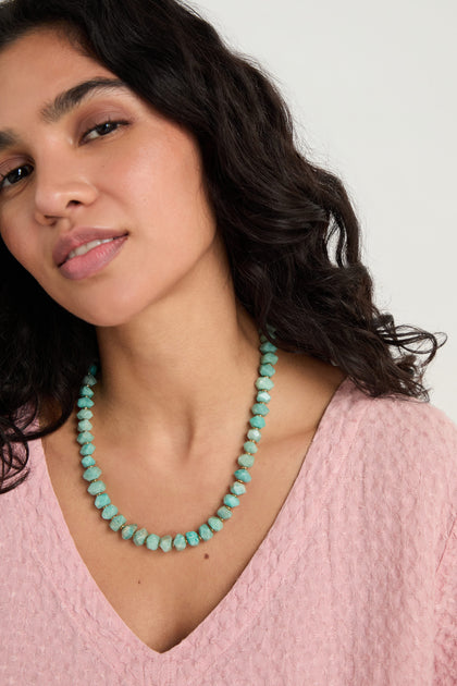 A woman with wavy dark hair wears a textured pink top and the Amazonite Bead Necklace, featuring a gold toggle clasp, as she looks slightly to the side against a plain light background.