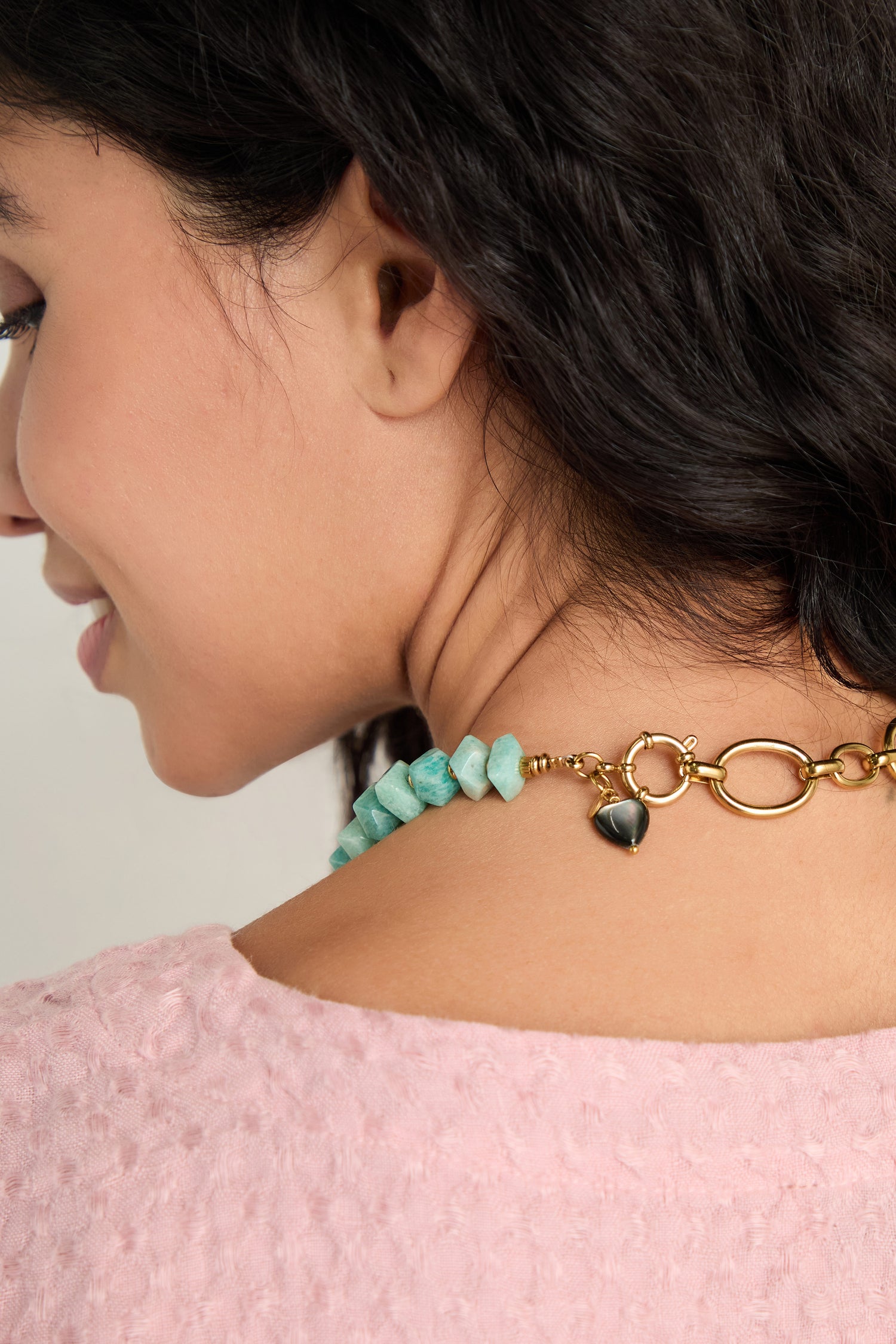 A woman with curly dark hair, shown from the side and back, wears the Amazonite Bead Necklace—chunky blue-green beads with a gold toggle clasp and black charm—with a pink textured top.