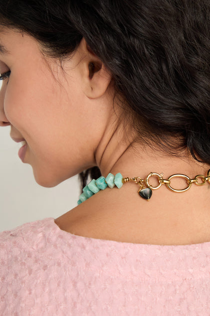 A woman with curly dark hair, shown from the side and back, wears the Amazonite Bead Necklace—chunky blue-green beads with a gold toggle clasp and black charm—with a pink textured top.
