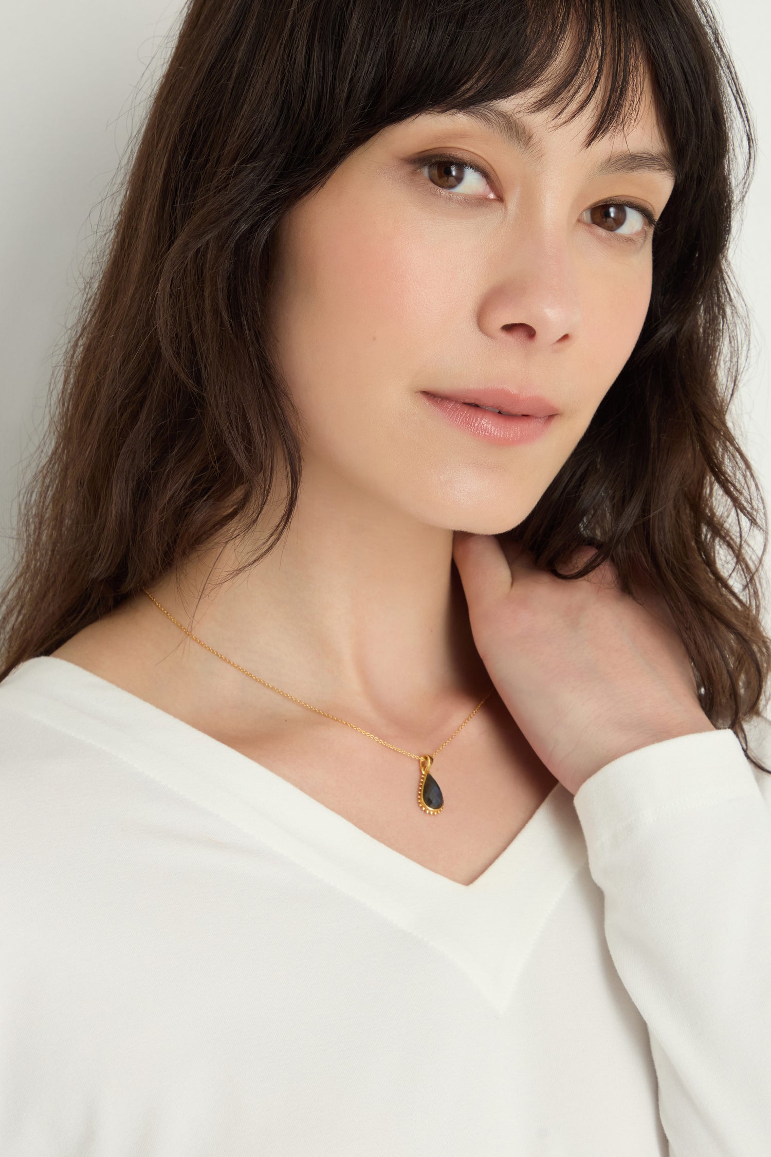 A woman with long brown hair wears a white top and the Gold Plated Labradorite Teardrop Necklace, touching her neck and looking at the camera.