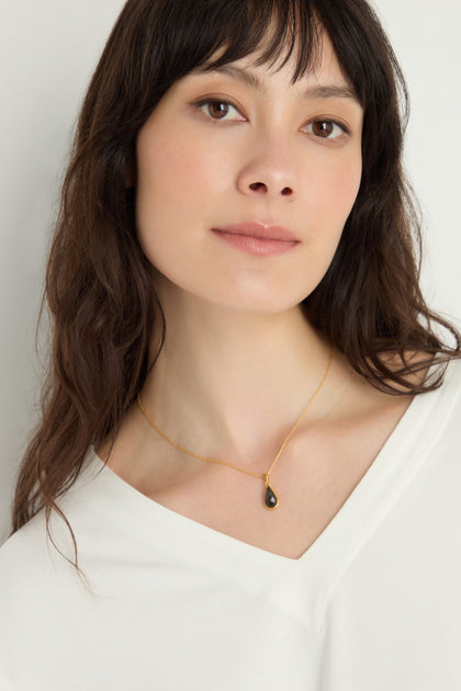 Woman with long brown hair wears a white top and a Gold Plated Labradorite Teardrop Necklace, featuring a small faceted labradorite pendant, as she looks at the camera against a plain background.
