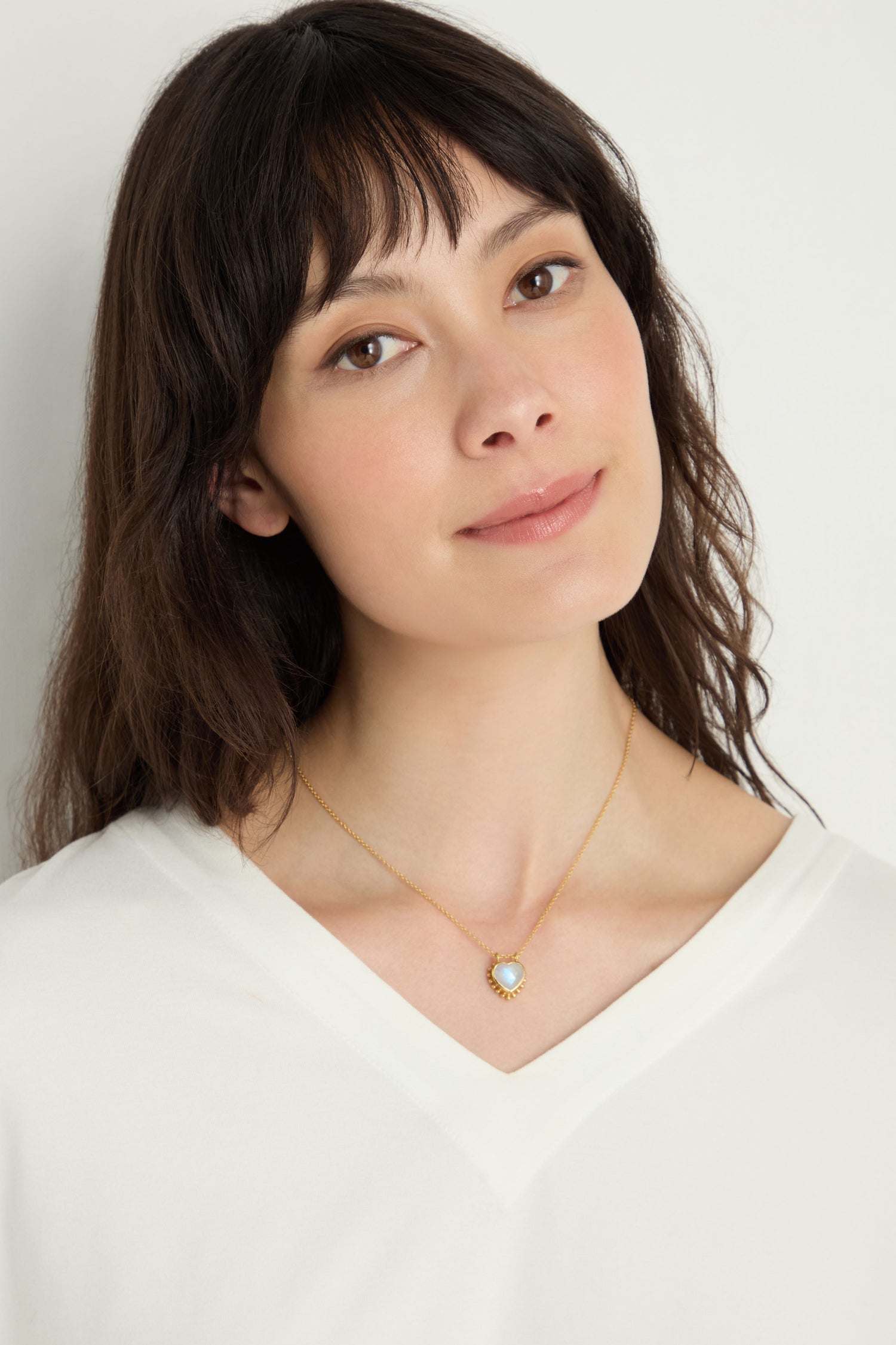 A woman with medium-length dark hair wears a white V-neck top and a Gold Plated White Moonstone Heart Necklace, featuring a heart-shaped pendant with a white moonstone, standing against a neutral background.