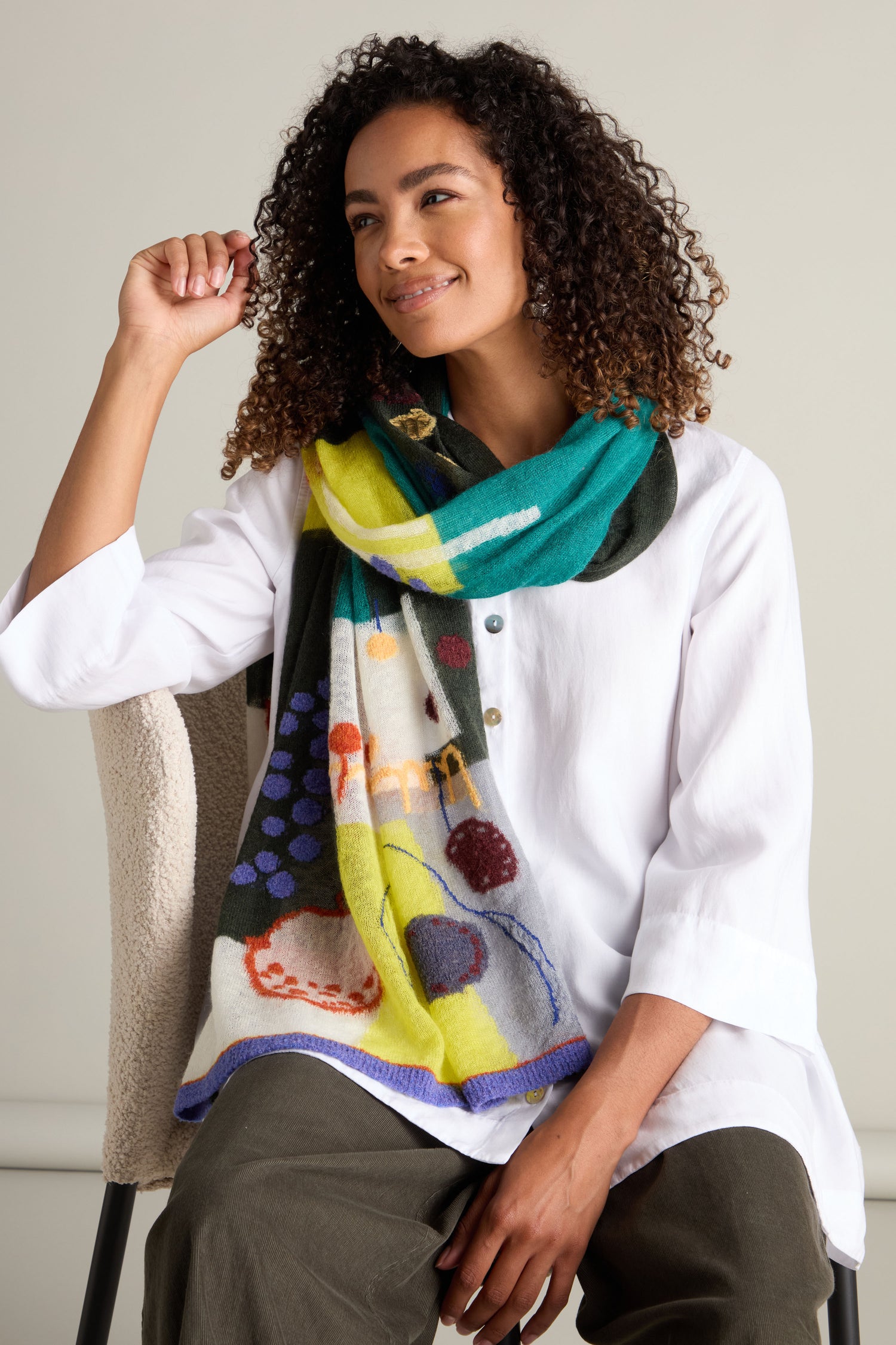 A person with curly hair wearing a white shirt and an Abstract Mohair Shawl sits on a chair, smiling and looking slightly to the right.