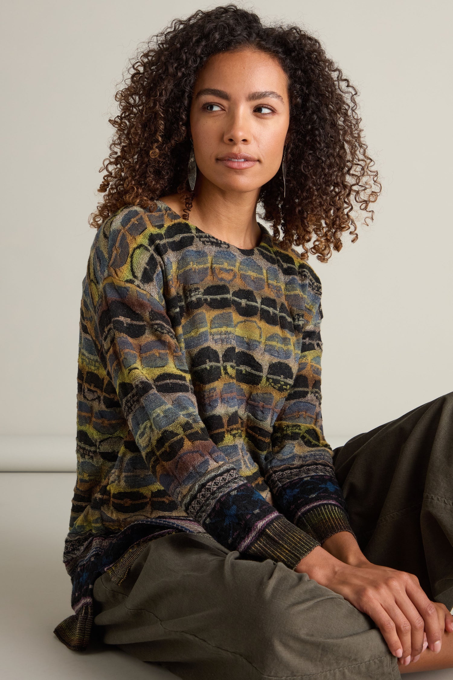 A person with curly hair wears a Round Neck Pullover and green pants, sitting on the floor against a neutral background, looking off to the side.