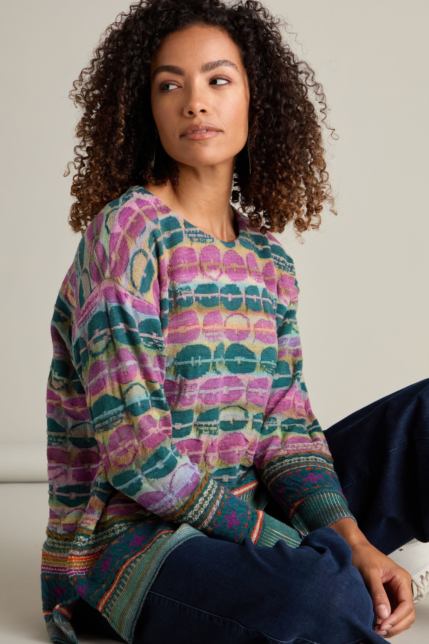 A woman with curly hair is wearing a Round Neck Pullover, featuring an intarsia knit with colorful patterns, paired with dark blue pants. She is seated and looking to the side, highlighting the artisanal excellence of the attire.