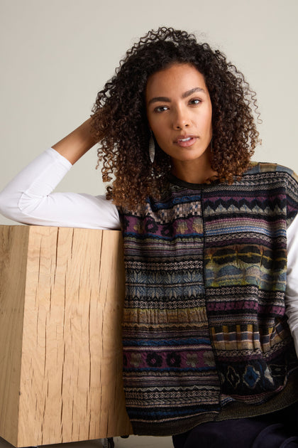 A woman with curly hair, wearing a Round Neck Tabard over a long-sleeved white shirt, leans against a wooden block.