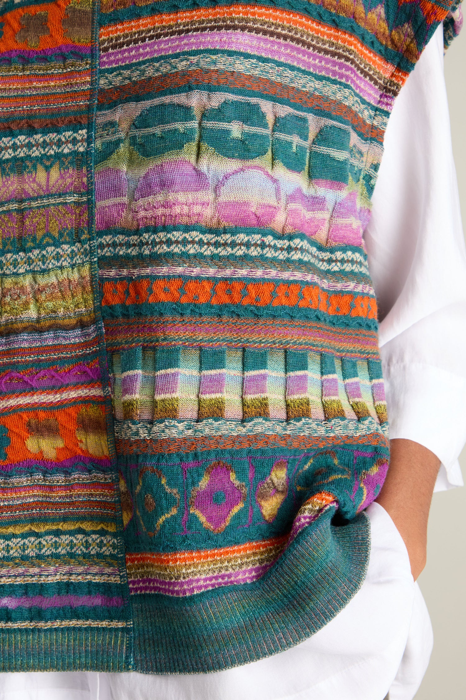 A person stands with one hand in their pocket, wearing a white shirt and a colorful, patterned Round Neck Tabard by M.&Kyoko, featuring stripes and geometric designs.