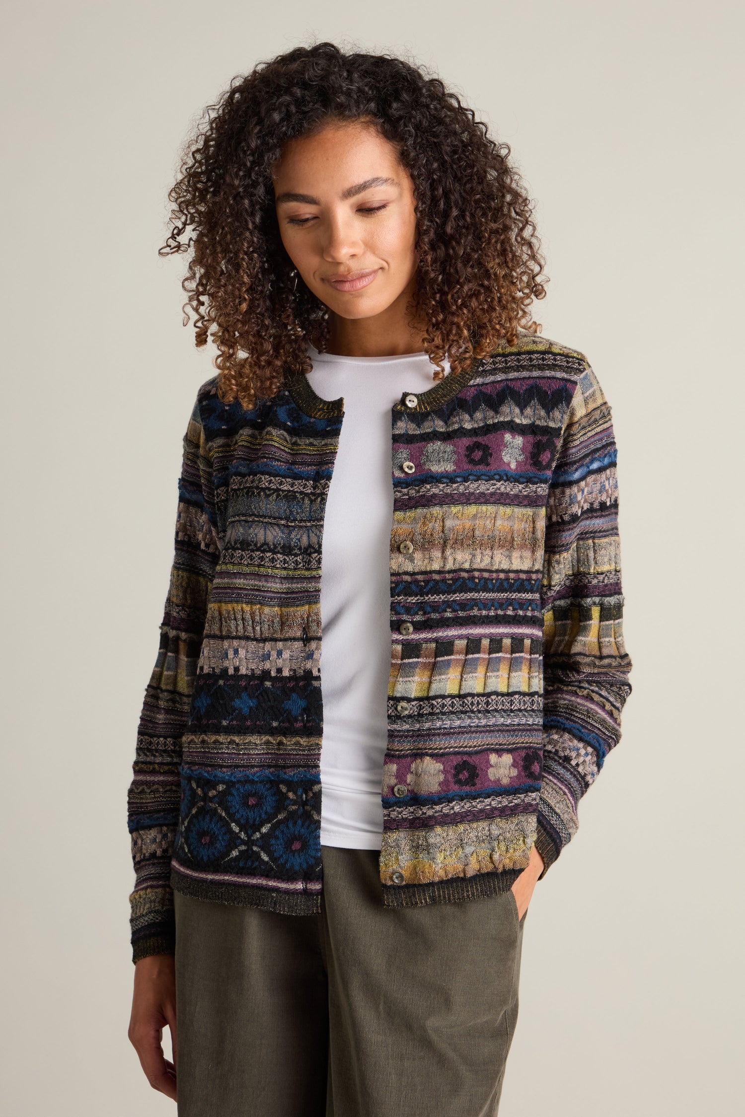 A woman with curly hair wears a round neck button cardigan with an intarsia knit multicolored pattern over a white shirt, posing with one hand in her pocket and looking down.