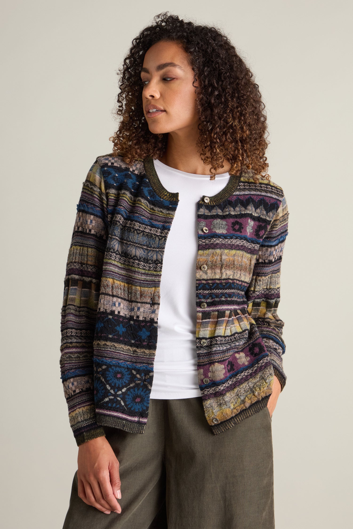 A person with curly hair is wearing a Round Neck Button Cardigan, characterized by its unique patterns, layered over a white top and paired with olive-green pants, posing against a neutral background.