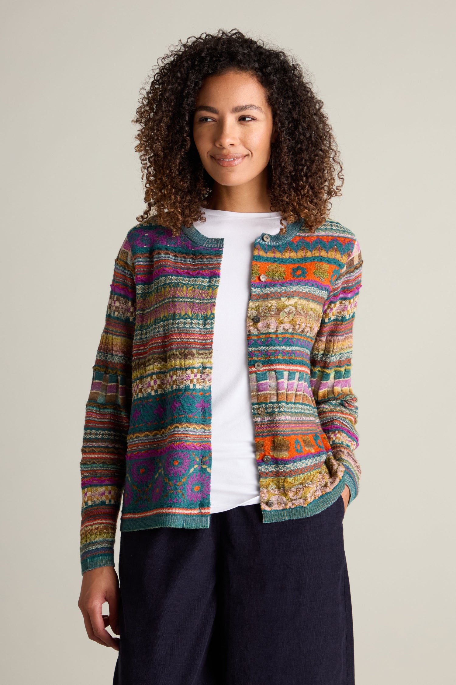 A person with curly hair is wearing the Round Neck Button Cardigan, featuring a colorful intarsia knit design, over a white shirt and dark pants, standing against a plain background.