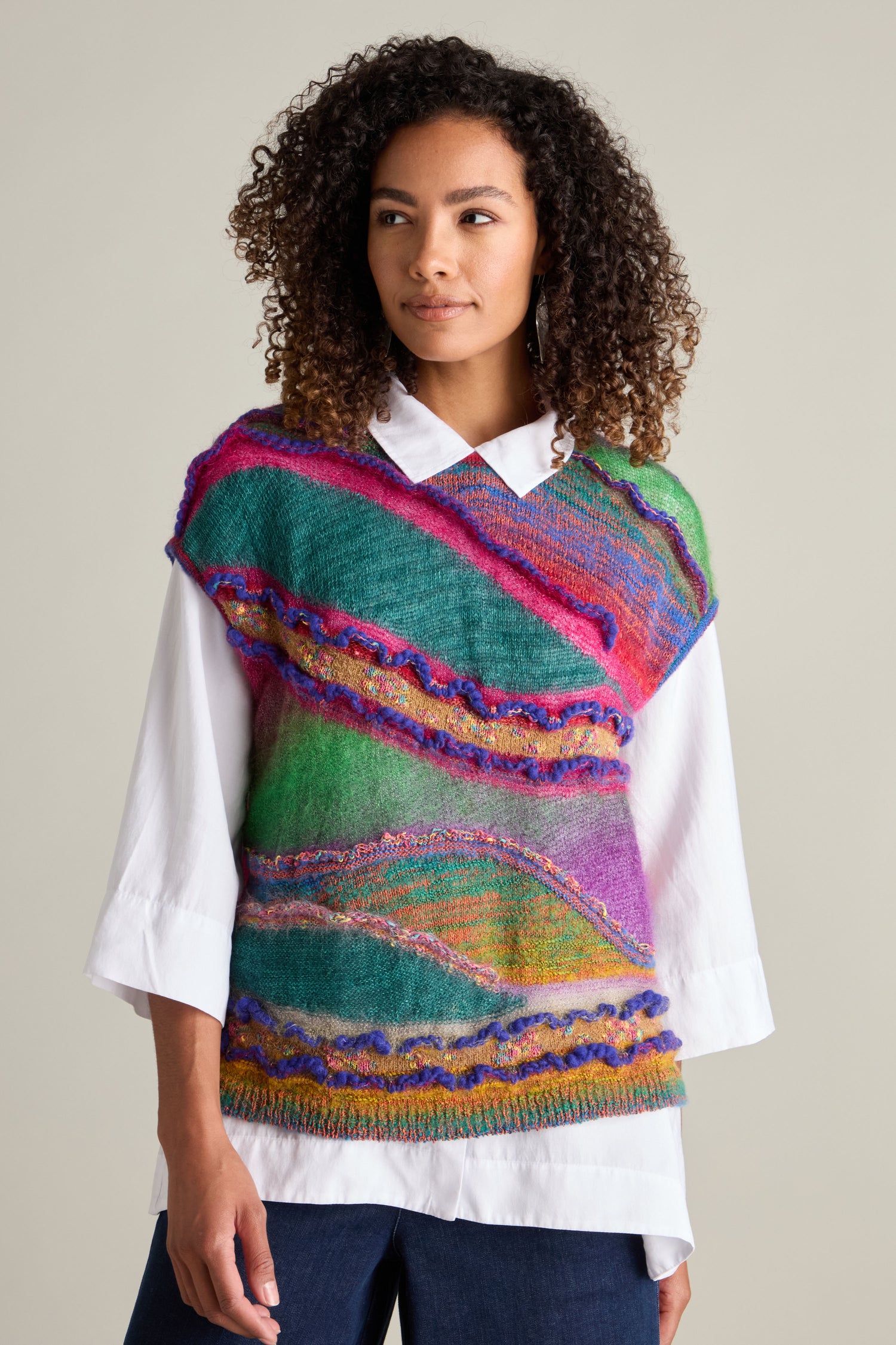 A person with curly hair wears a vibrant Stitch Detail Tabard over a white shirt, looking to the side.