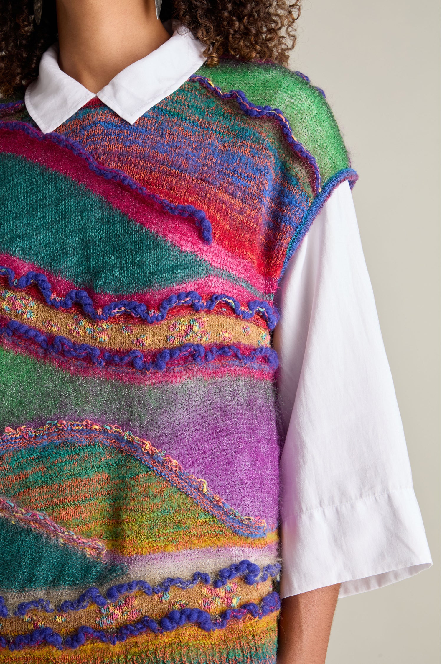 A person wearing a white collared shirt under the Stitch Detail Tabard in vibrant hues, showcasing the charm of modern knitwear.