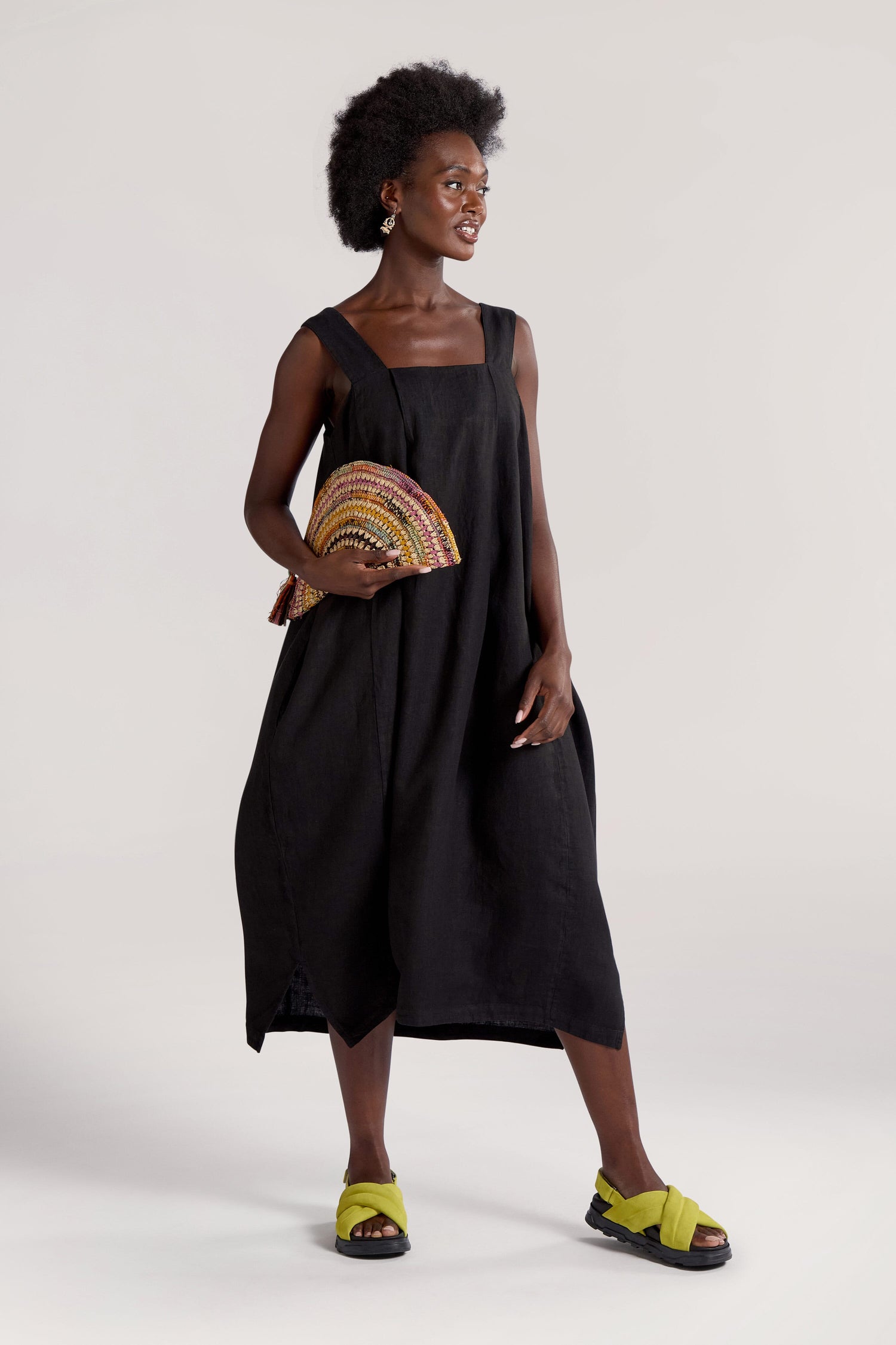 A person in a stylish Square Neck Bubble Linen Dress, ideal for any summer rotation, holds a colorful semi-circle clutch bag. They wear green-yellow sandals and stand against a plain background.