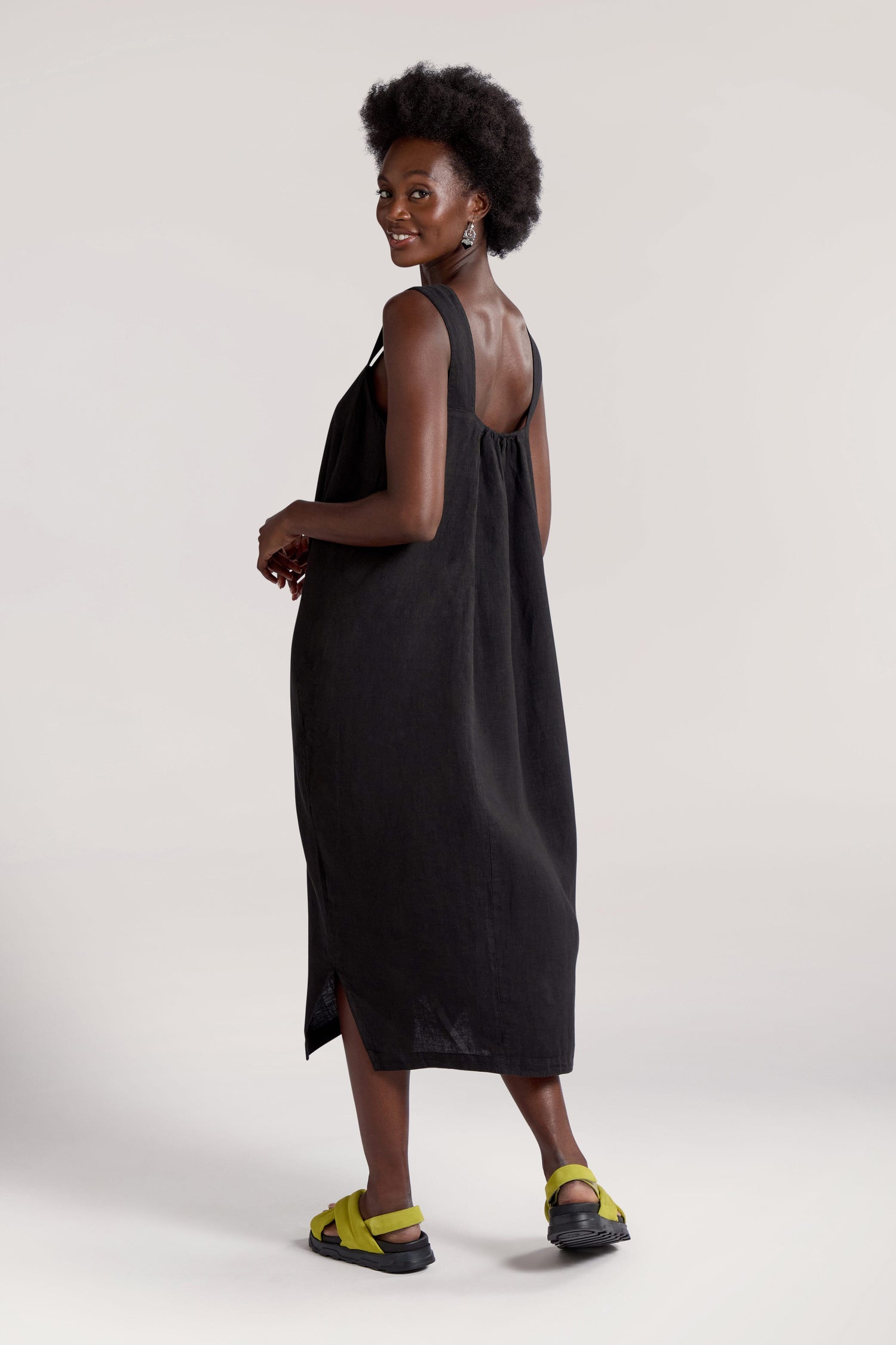 A person wearing a sleeveless black Square Neck Bubble Linen Dress and green sandals stands and smiles, looking over their shoulder against a neutral background – the perfect addition to your summer rotation.