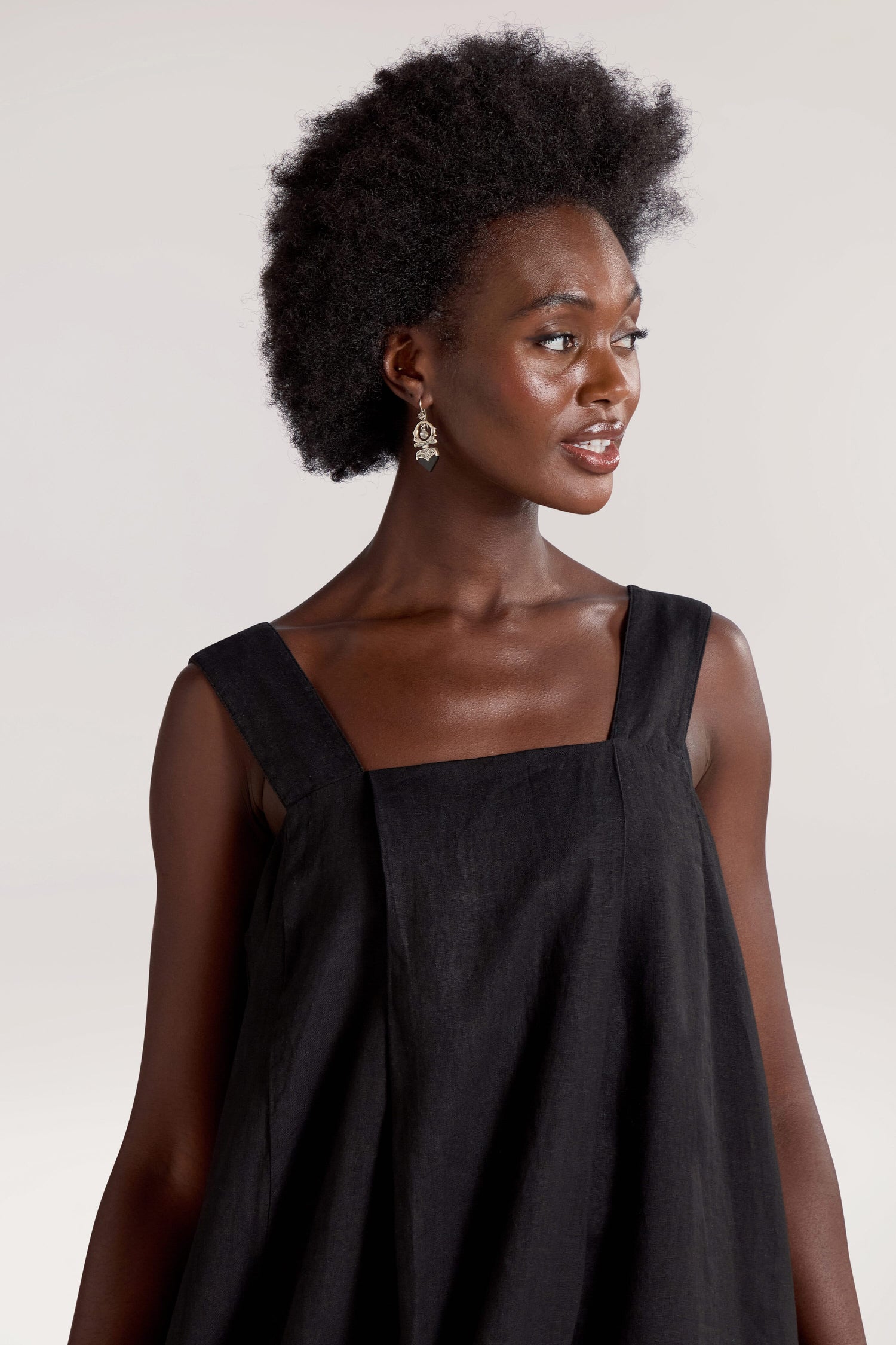 A woman with an afro hairstyle wearing a Square Neck Bubble Linen Dress and earrings looks to her right.