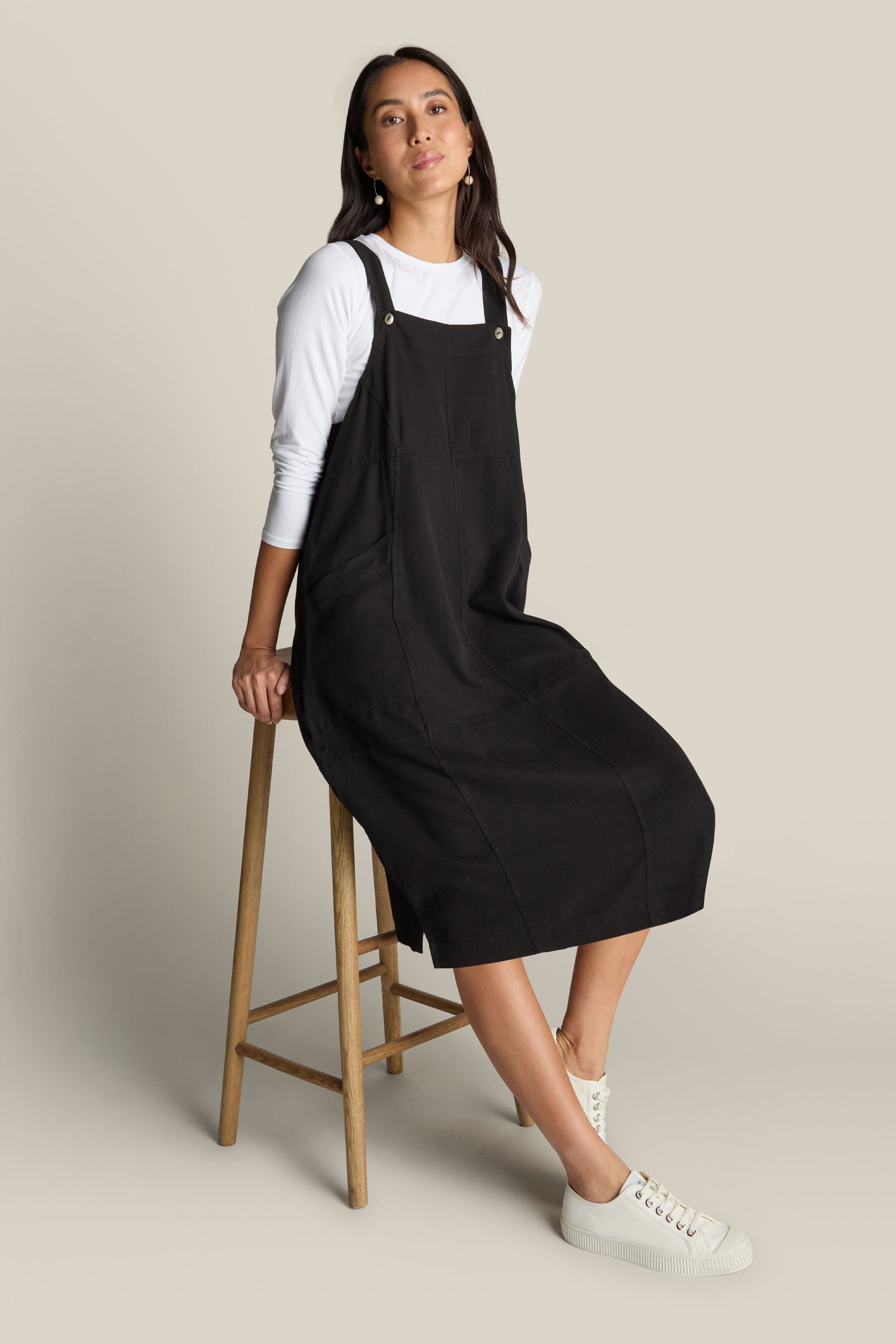 A woman is sitting on a wooden stool, wearing a Fluid Twill Pinafore Dress paired with a white long-sleeve shirt and white sneakers. She exudes bohemian charm as she gazes slightly to the side.