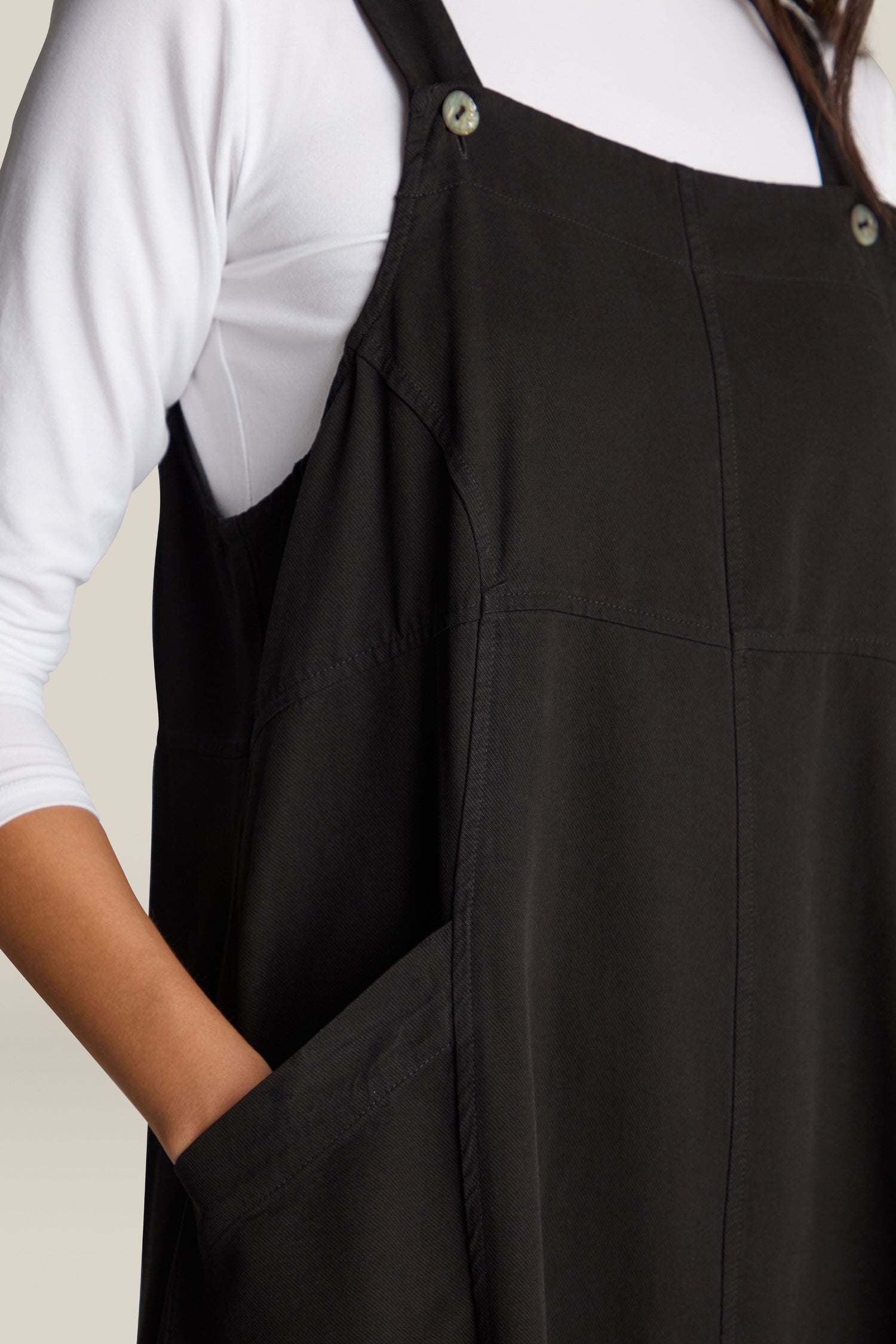 A person wearing the Fluid Twill Pinafore Dress over a white long-sleeve shirt is depicted with one hand in the dress pocket, radiating bohemian charm.