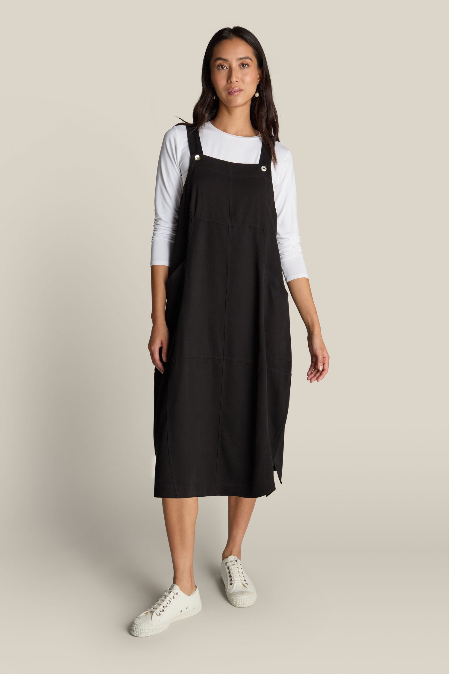 A woman stands against a plain background, showcasing bohemian charm in a knee-length Fluid Twill Pinafore Dress paired with a white long-sleeve shirt and white sneakers.