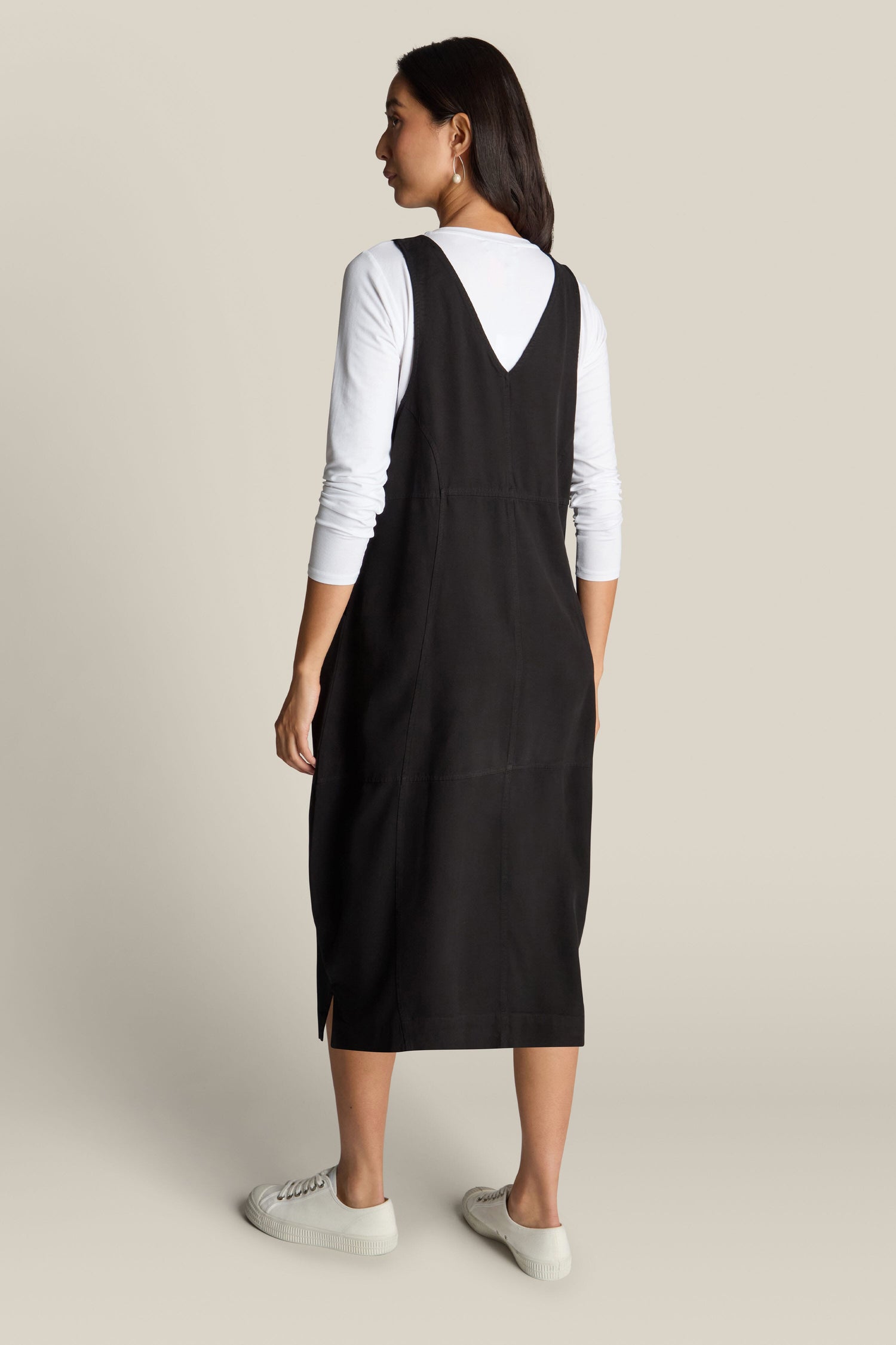 A woman is standing sideways, showcasing the luxurious Fluid Twill Pinafore Dress in black, elegantly layered over a white long-sleeve shirt and paired with white sneakers.