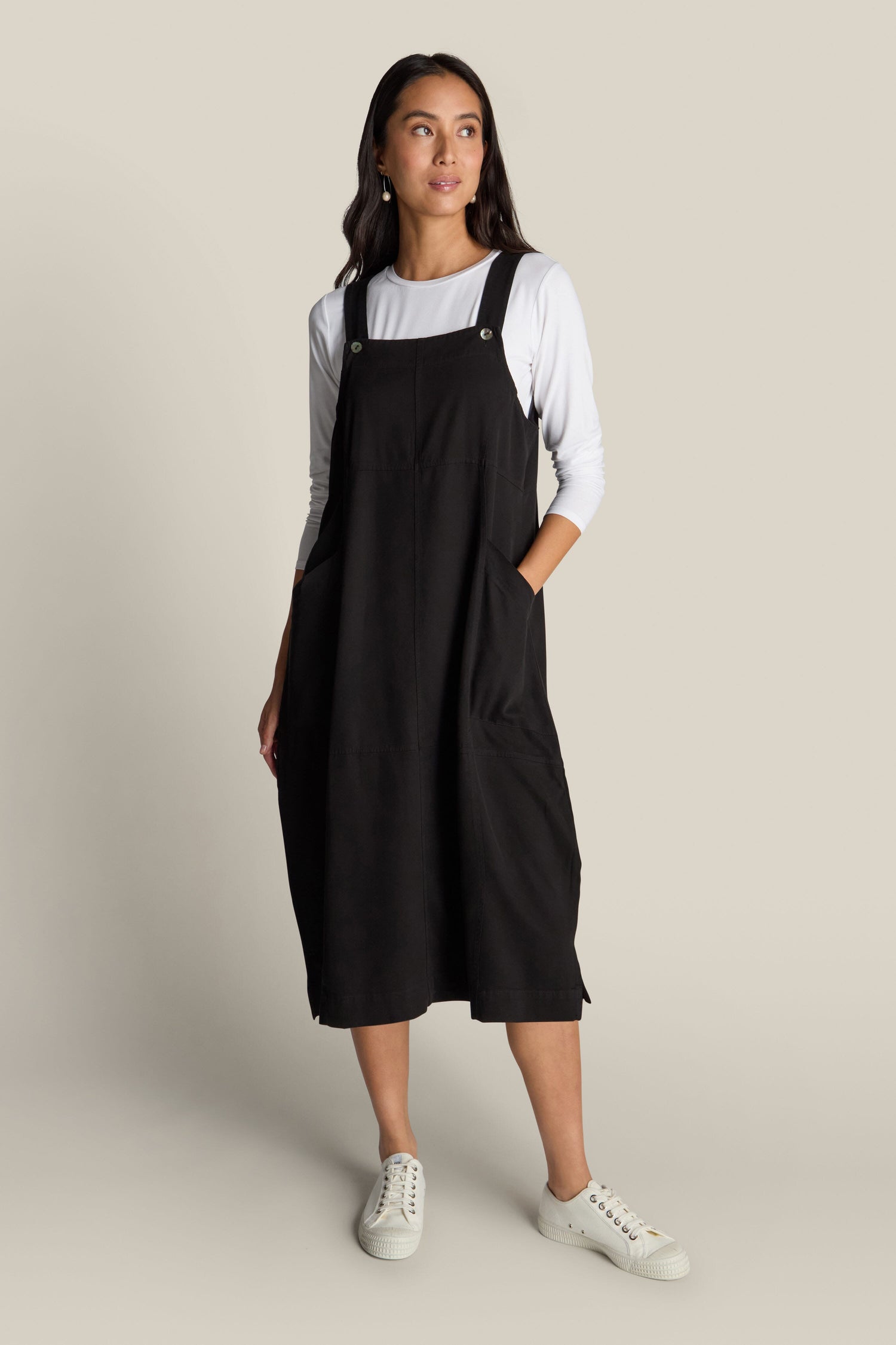 A woman is pictured against a neutral backdrop, dressed in a Fluid Twill Pinafore Dress layered over a white long sleeve shirt and paired with white sneakers. With her hands tucked into the dress's pockets, she exudes effortless bohemian charm.