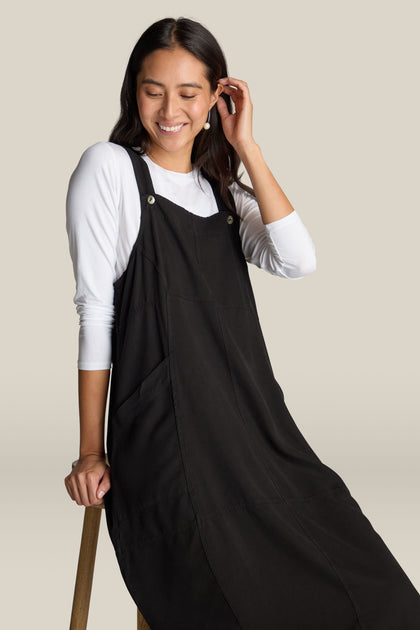 A woman with long hair, wearing a luxurious Fluid Twill Pinafore Dress over a white long-sleeve shirt, sits on a stool and smiles while touching her ear.