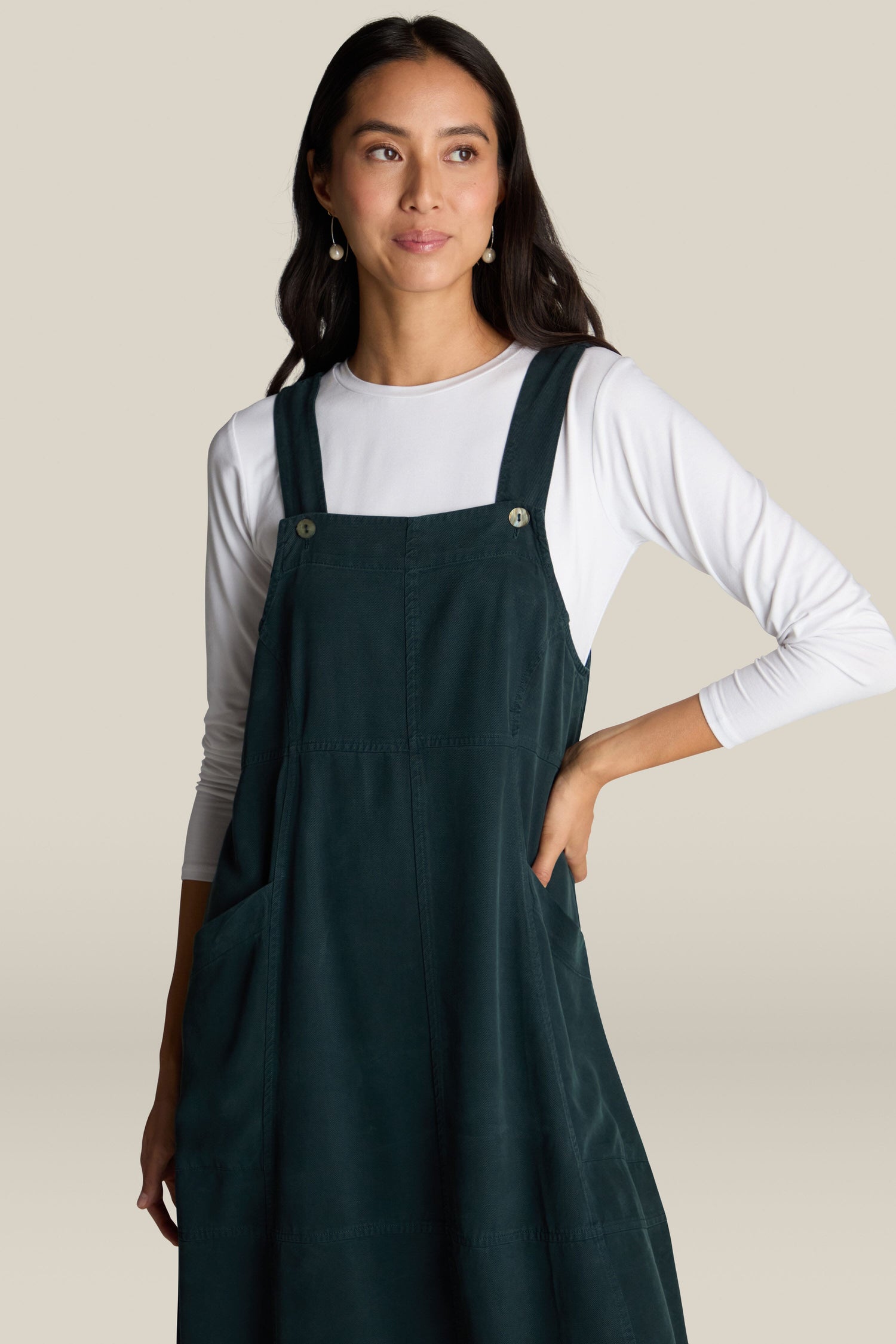 A woman with long dark hair is wearing the luxurious Fluid Twill Pinafore Dress over a white long-sleeve shirt, exuding bohemian charm. She has one hand on her hip and is looking slightly to the side.