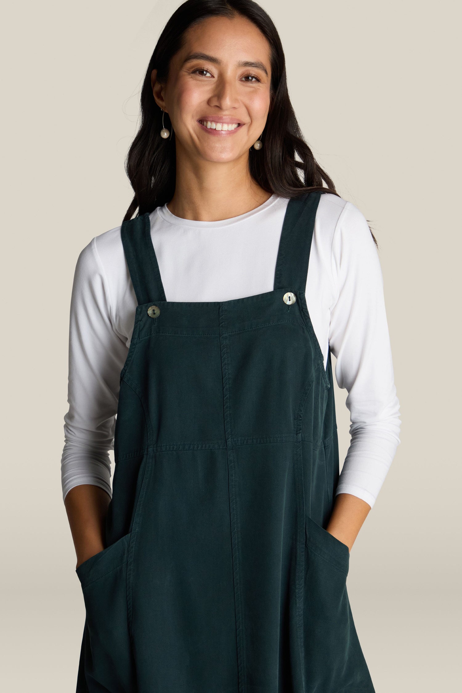 A woman with long dark hair smiles while wearing a white long-sleeve shirt beneath a dark green Fluid Twill Pinafore Dress featuring pockets, radiating bohemian charm. She stands against a simple, light-colored backdrop.