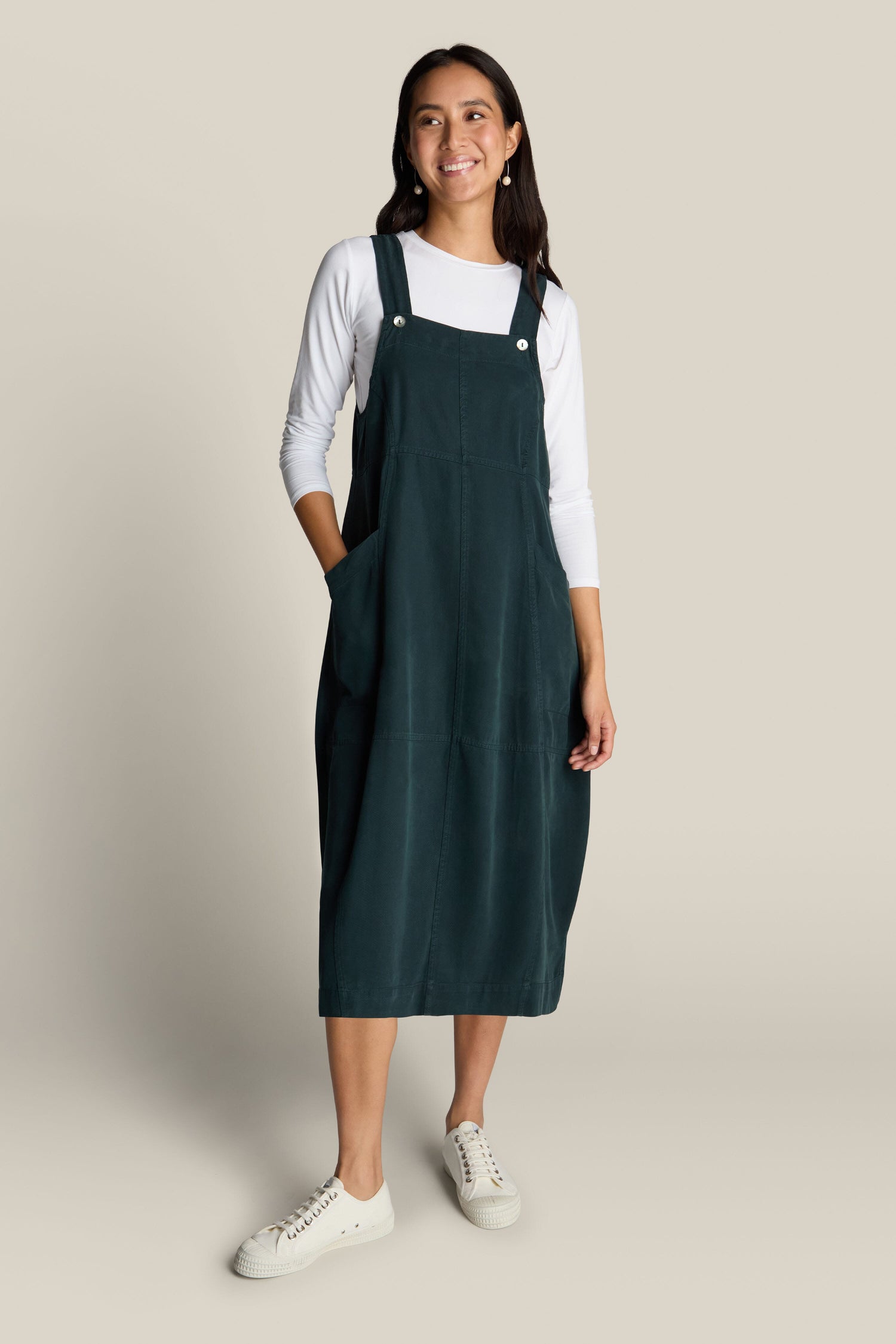 A woman is standing and smiling, wearing a luxurious Fluid Twill Pinafore Dress in dark green over a white long-sleeve shirt with white sneakers. She has her hands in the pockets of the dress, exuding bohemian charm against a plain beige background.