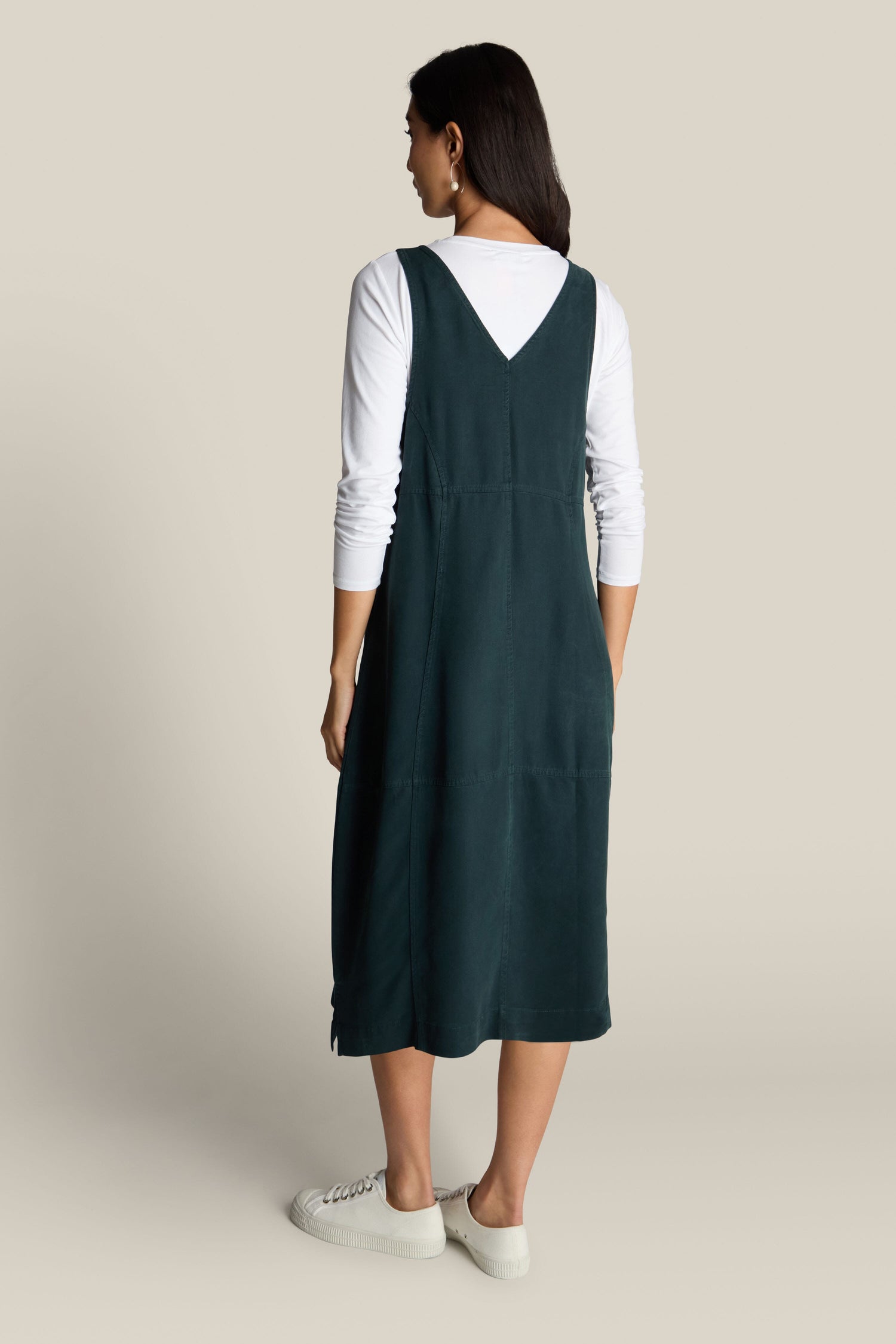 A woman stands turned away, radiating bohemian charm in a Fluid Twill Pinafore Dress, layered over a white long-sleeve shirt and complemented by white sneakers.