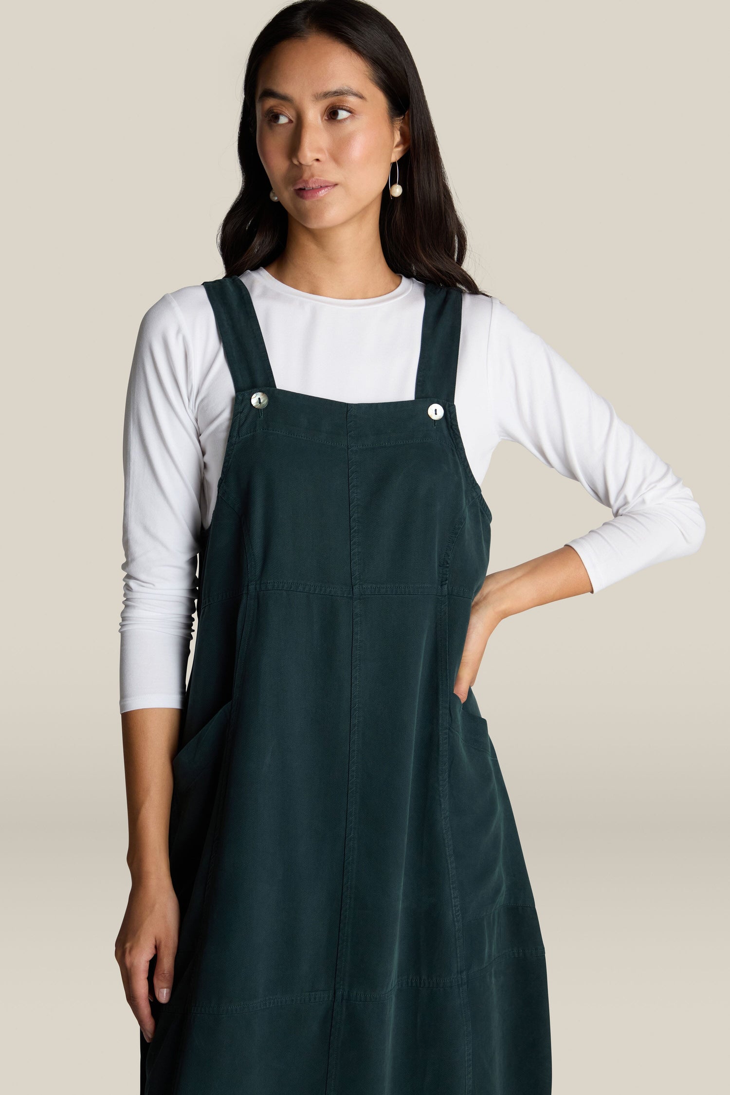 A person stands against a plain background wearing a white long-sleeve shirt under a dark green Fluid Twill Pinafore Dress, effortlessly exuding bohemian charm.