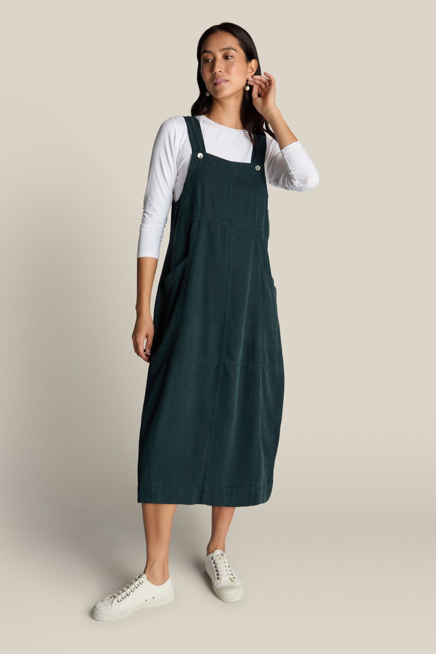 A person is wearing the Fluid Twill Pinafore Dress in dark green over a white long-sleeve shirt, paired with white sneakers against a plain light background.