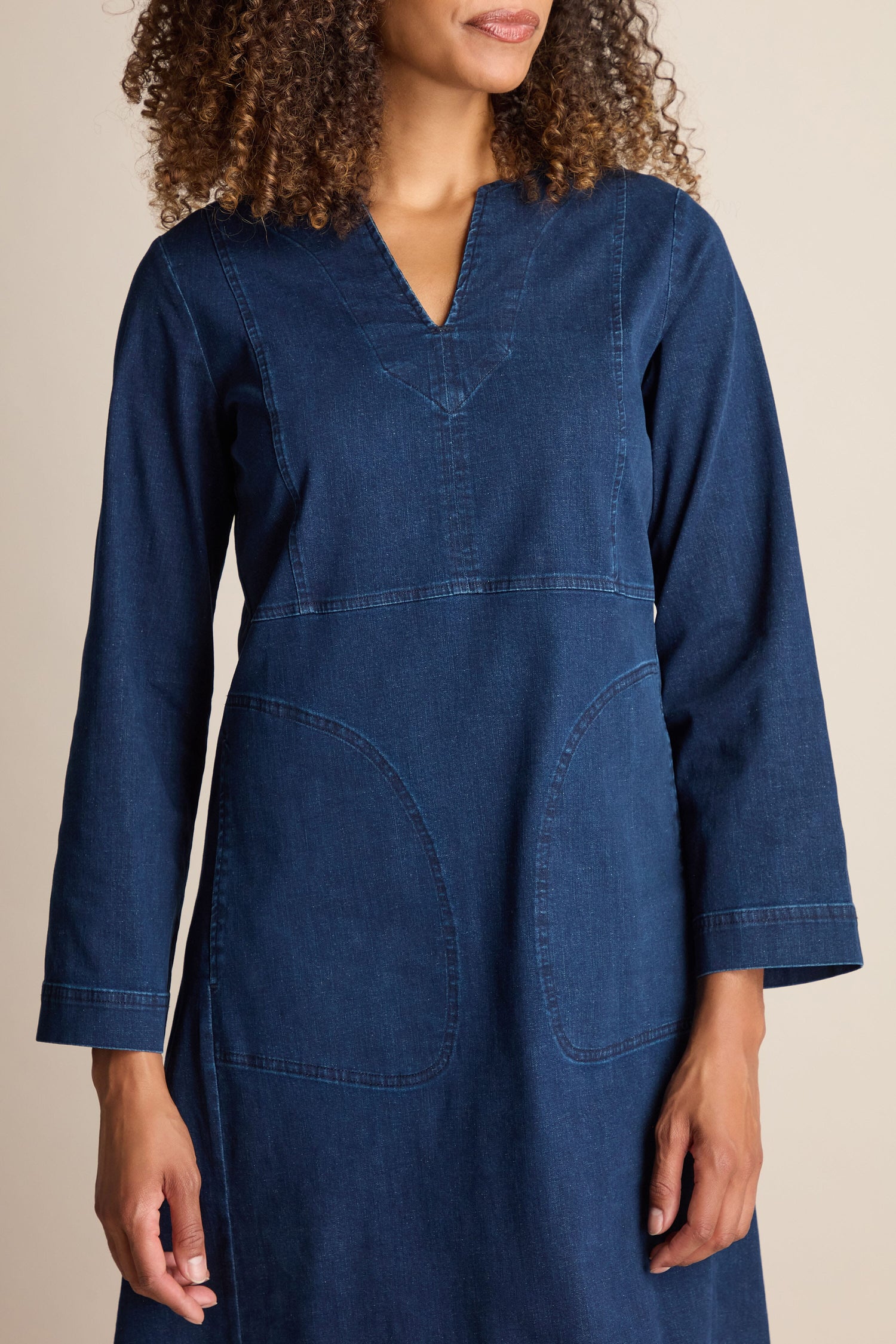 A person with curly hair wears a long-sleeve Stretch Denim Pocket Dress in dark blue, which features an A-line silhouette, v-neckline, and front pockets.