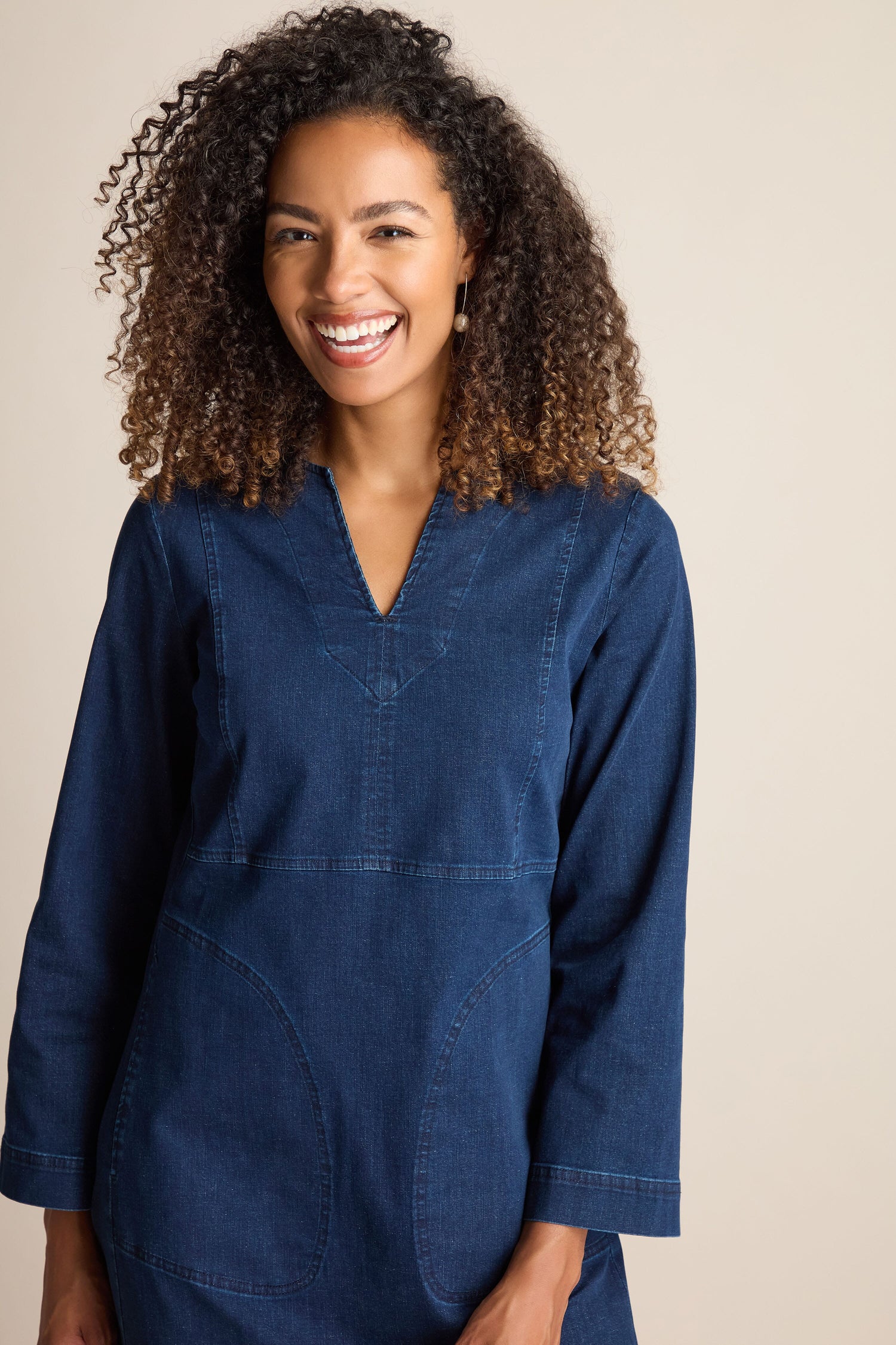 A person with curly hair smiles while wearing the Stretch Denim Pocket Dress in blue, featuring long sleeves and an A-line silhouette.