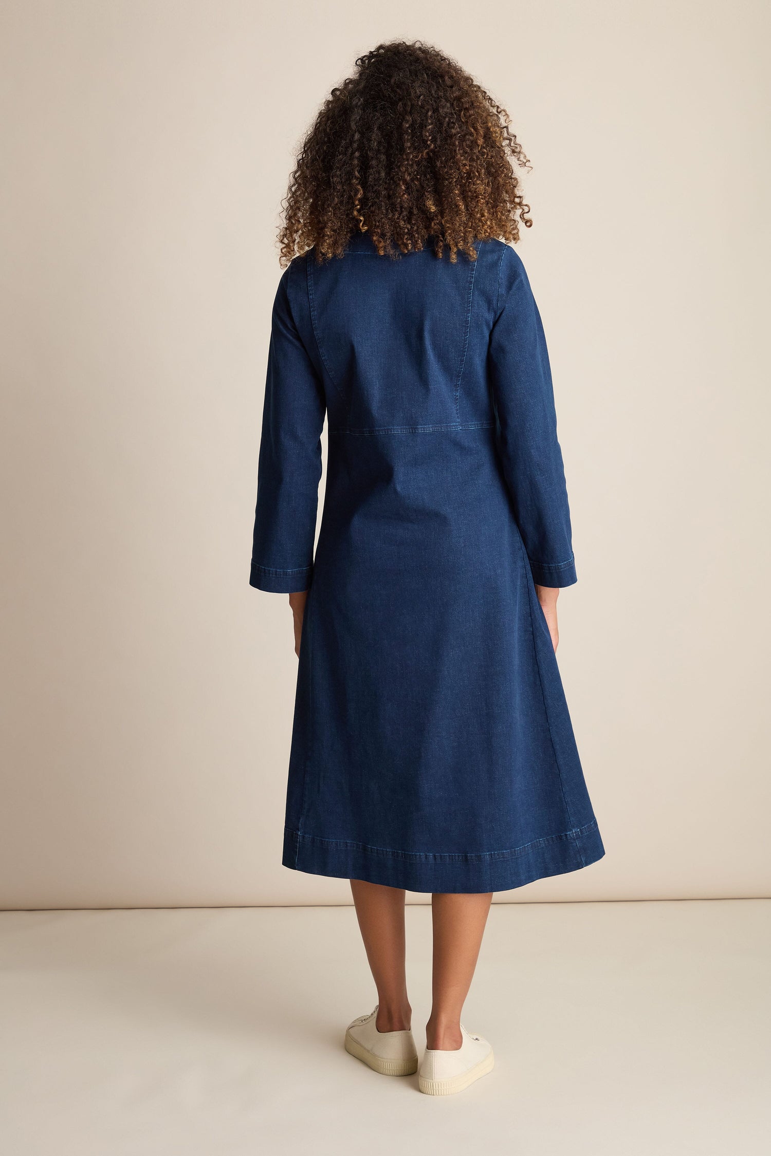 A person with curly hair is standing facing away, wearing a Stretch Denim Pocket Dress with an A-line silhouette, long sleeves, and knee-length cut in dark blue denim, paired with white shoes.