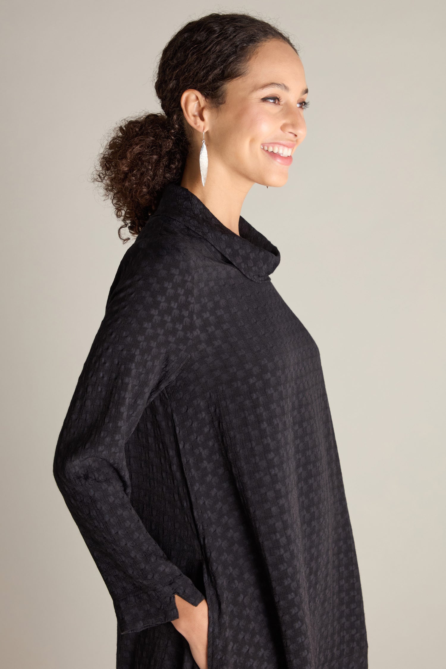 A woman with curly hair styled in a ponytail, wearing a black Small Square Jacquard Bubble Dress, smiles while looking to the side. She has one hand in her pocket and is also wearing large white earrings.