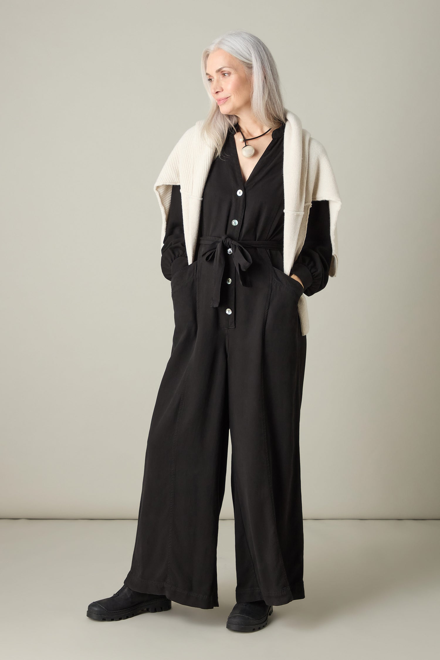 A woman with long gray hair stands against a plain background, wearing a Fluid Twill Jumpsuit in black, featuring a contemporary rounded neckline, and paired with a cream sweater. Her hands rest casually in her pockets, highlighting the jumpsuit's eco-friendly fabric and modern style.